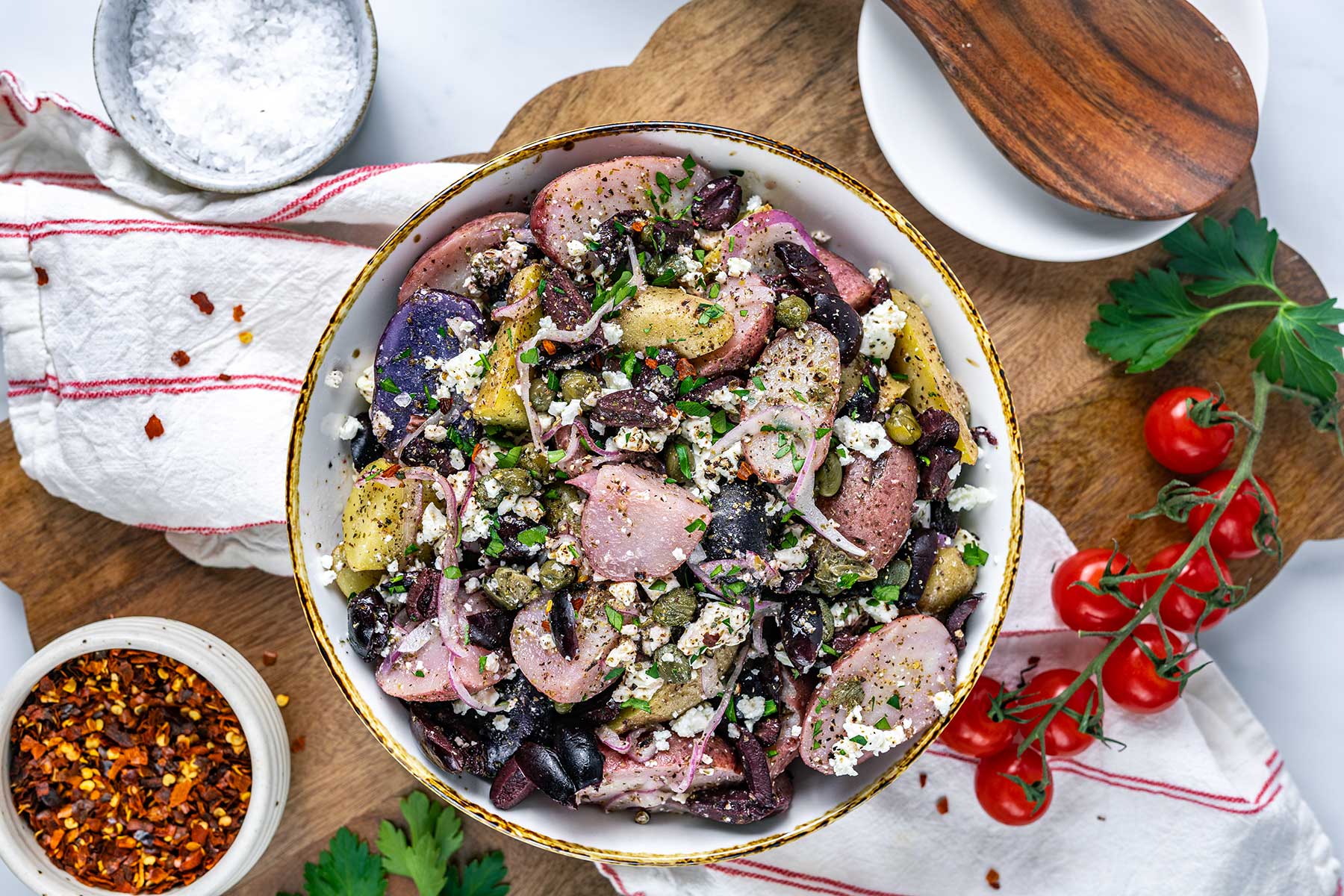 A bowl of potato salad with black beans, chopped herbs, and feta cheese sits on a wooden board, surrounded by cherry tomatoes, sautéed mushrooms for steak, a bowl of salt, red pepper flakes, and a white towel with red stripes.
