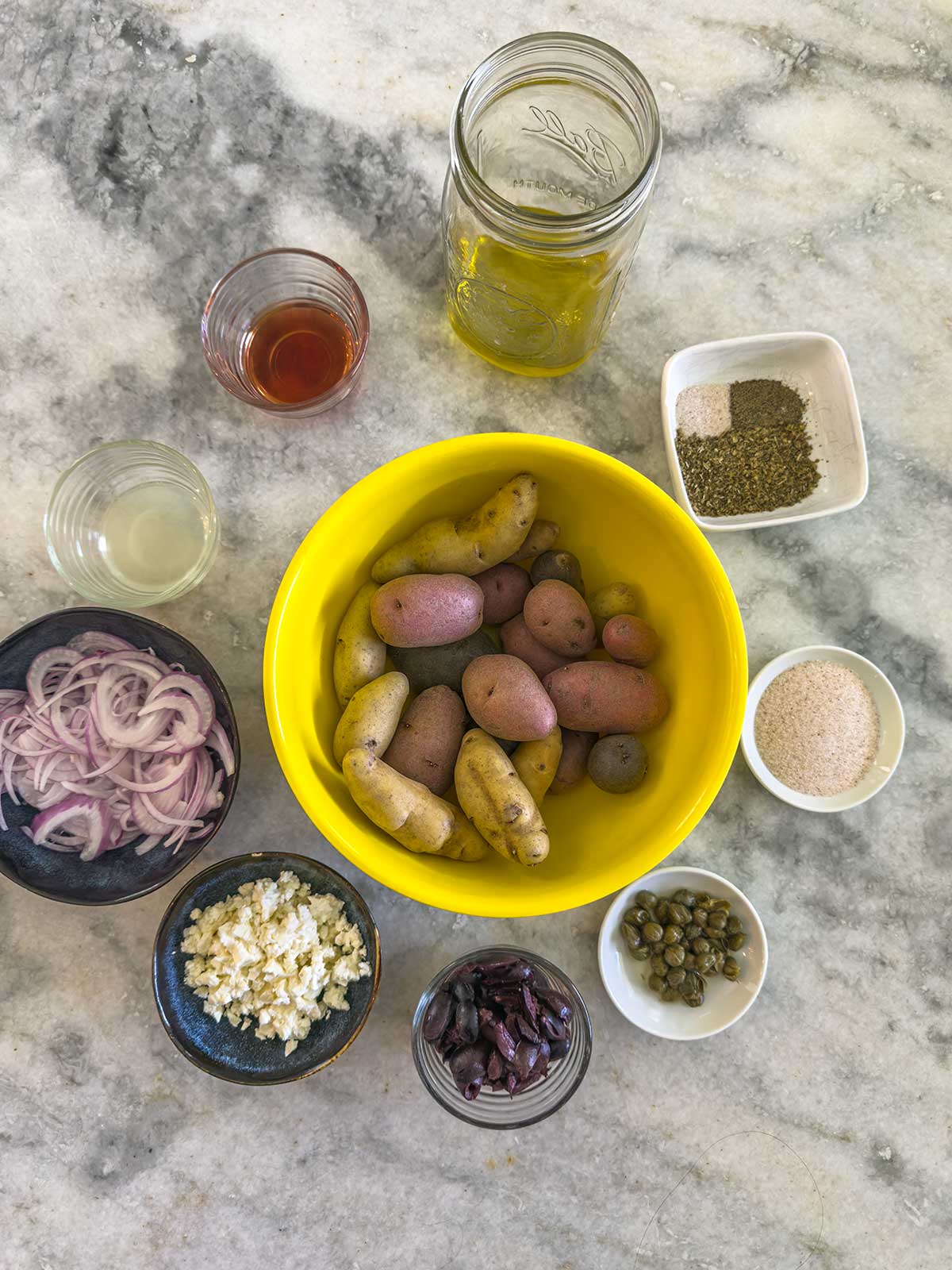 A yellow bowl of Greek potato salad made with fingerling potatoes is surrounded by small bowls of sliced red onion, feta cheese, chopped olives, capers, vinegar, lemon juice, olive oil, salt, and black pepper on a marble countertop.