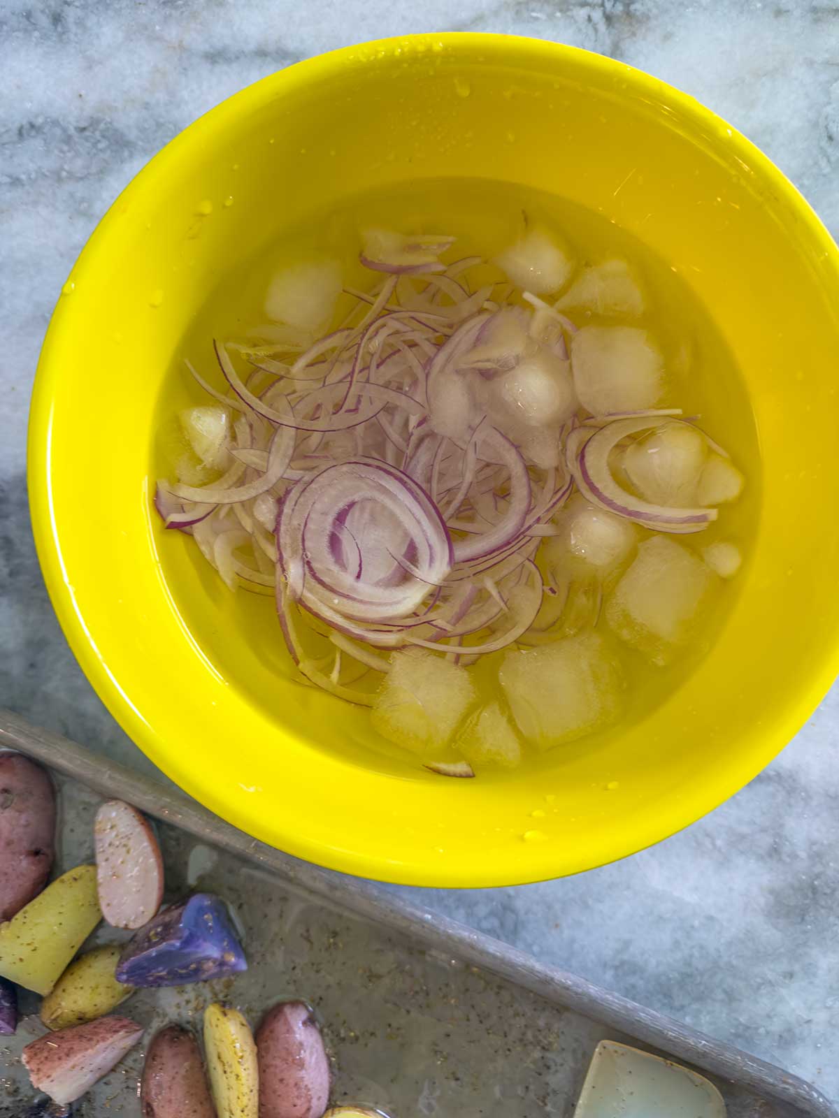 A yellow bowl filled with water, ice cubes, and thinly sliced red onions sits on a marble surface. Next to it, a tray with colorful cut potatoes—perfect for crafting a refreshing Greek potato salad—is partially visible.
