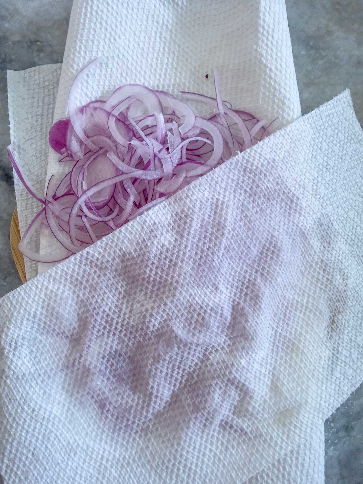 Thinly sliced red onions, ideal for greek potato salad, rest on a paper towel and are partially covered by another sheet on a light-colored surface. The paper towels appear slightly damp.