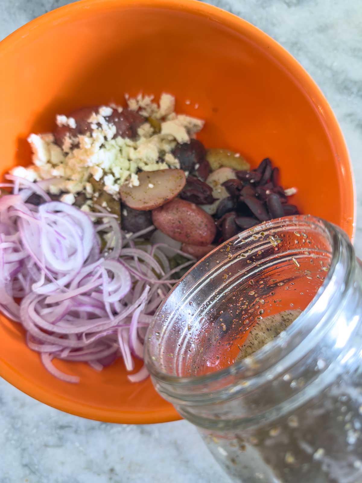 A jar of dressing is being poured into an orange bowl containing sliced red onions, kidney beans, chunked potatoes, and minced garlic on a light countertop—perfect for making a delicious Greek potato salad.