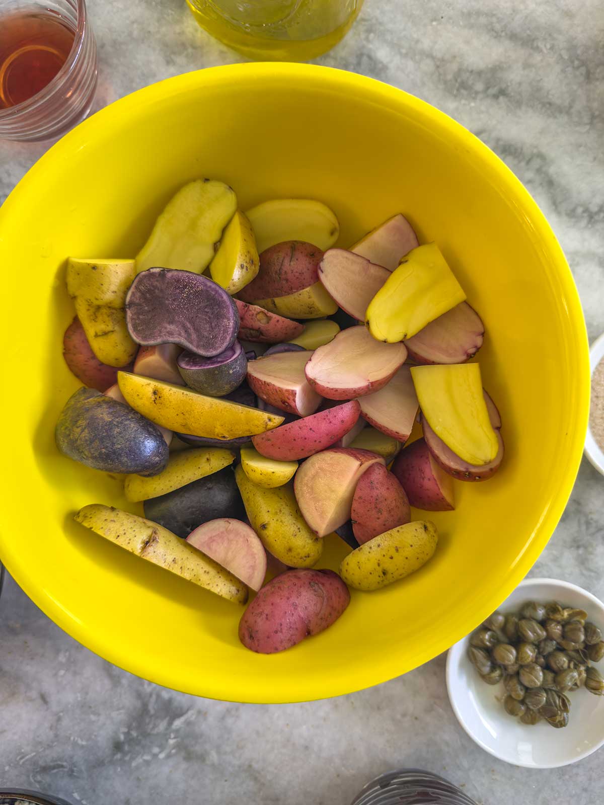 A yellow bowl filled with various types of sliced potatoes in purple, yellow, and red hues sits on a marble surface. Surrounding it are small bowls with capers and other ingredients for a vibrant Greek potato salad.