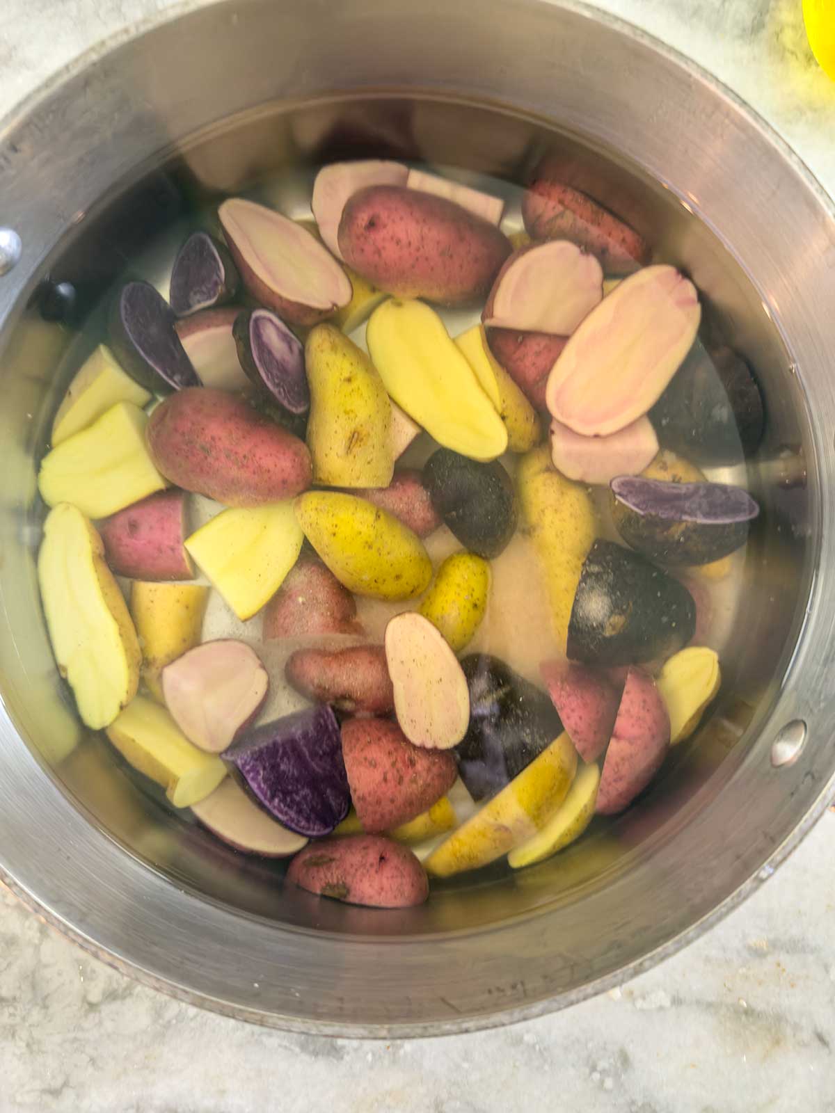 A stainless steel bowl filled with water and a mix of halved and whole small potatoes in various colors, including red, yellow, and purple, sits on a marble countertop—perfect for making a vibrant Greek potato salad.