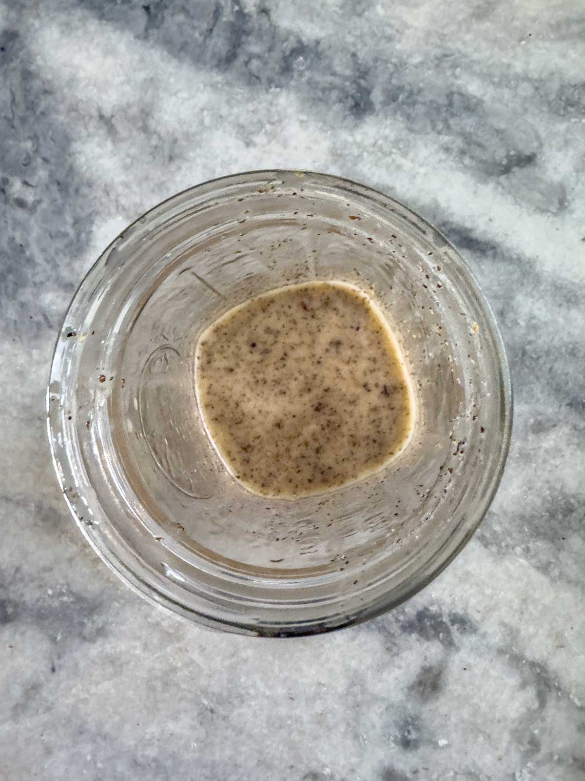 A glass jar containing a small amount of liquid and dark specks, reminiscent of greek potato salad seasonings, sits on a gray and white marbled surface, viewed from above.