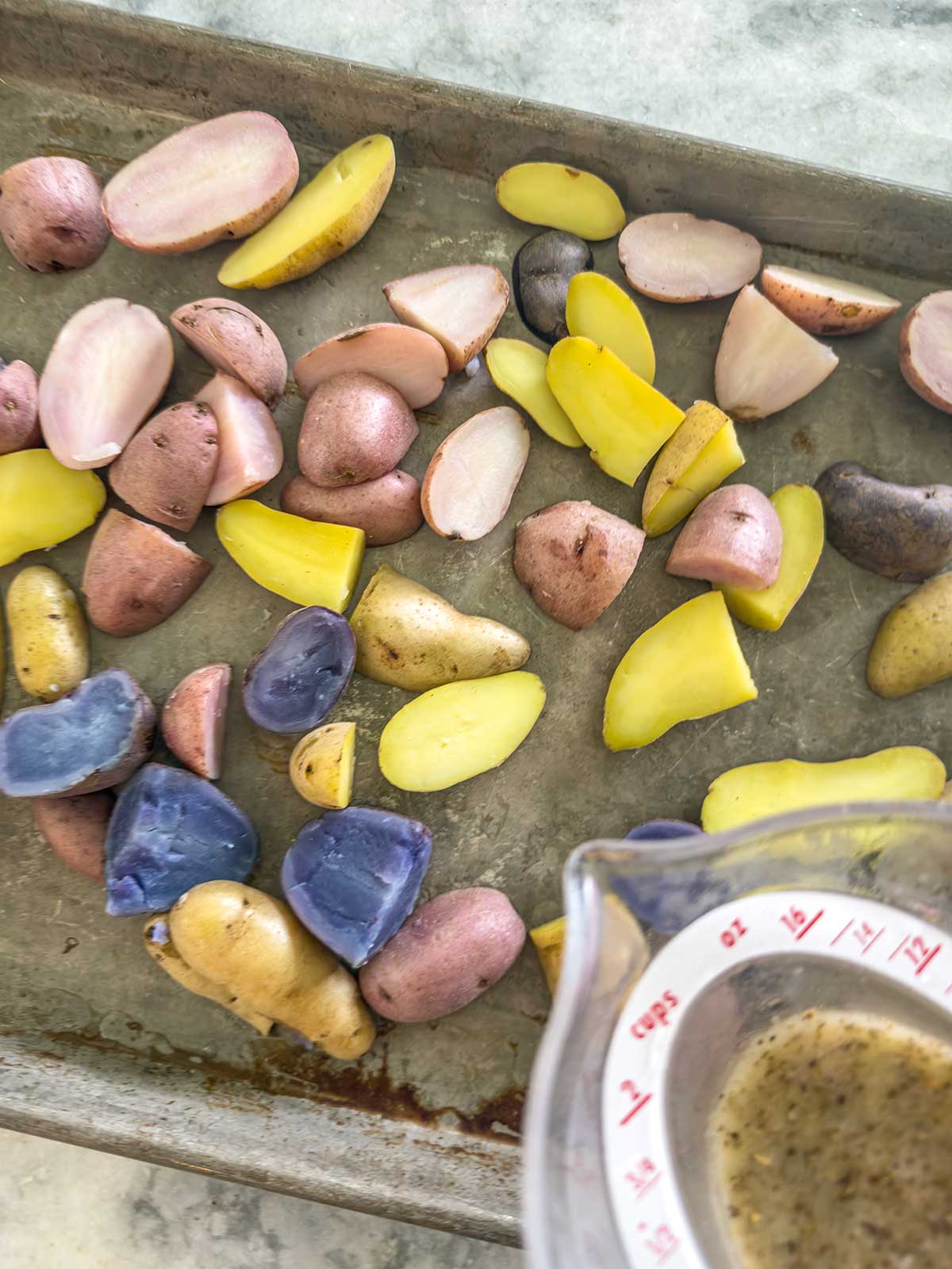 Sliced red, yellow, and purple baby potatoes are spread on a metal baking sheet. In the corner, a measuring cup with seasoned liquid for a greek potato salad is being poured onto the potatoes.