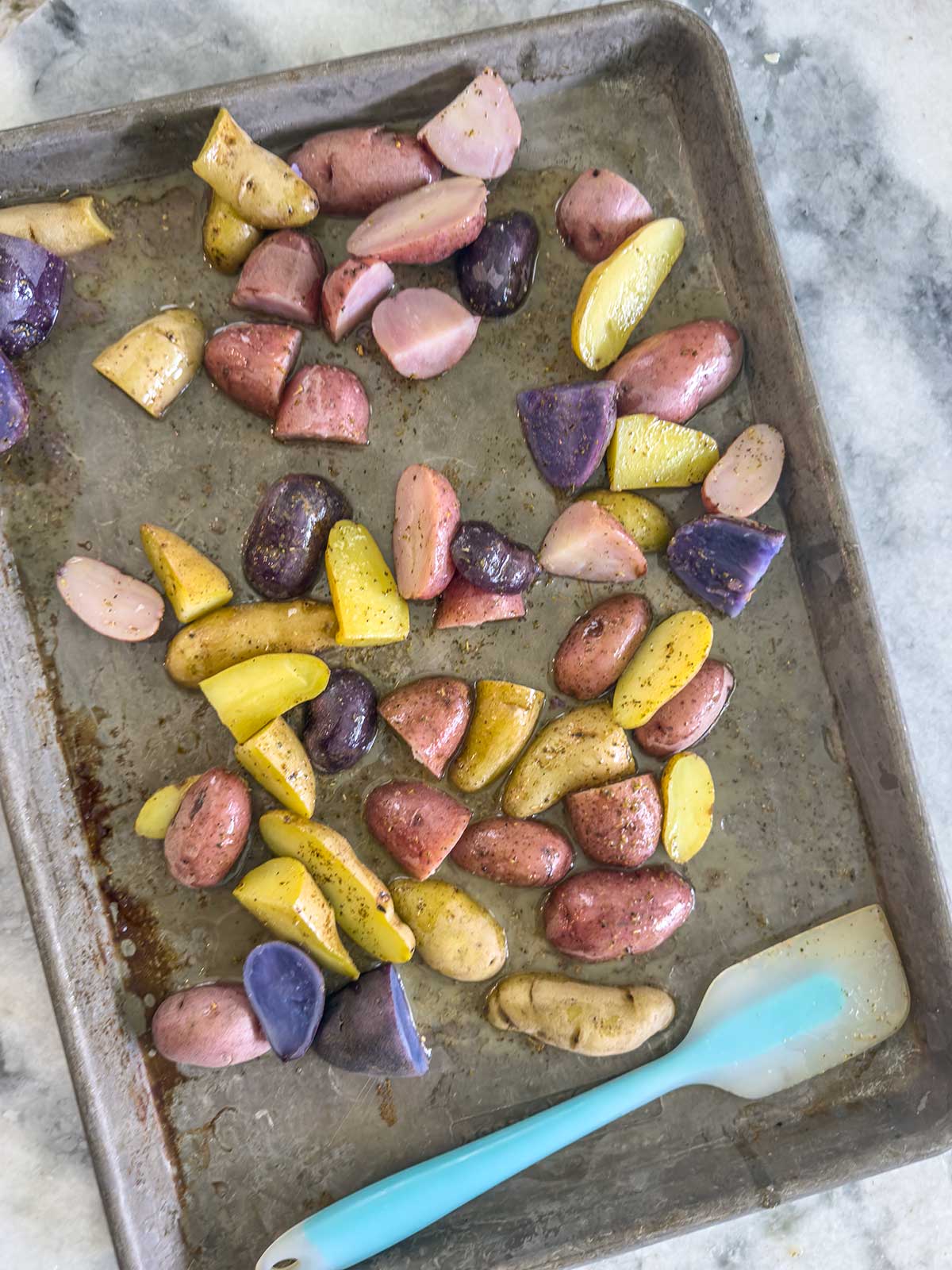 A baking sheet with seasoned, cut red, yellow, and purple potatoes—perfect for making greek potato salad. Some pieces are scattered, and a blue spatula rests at the bottom right on a light marble countertop.