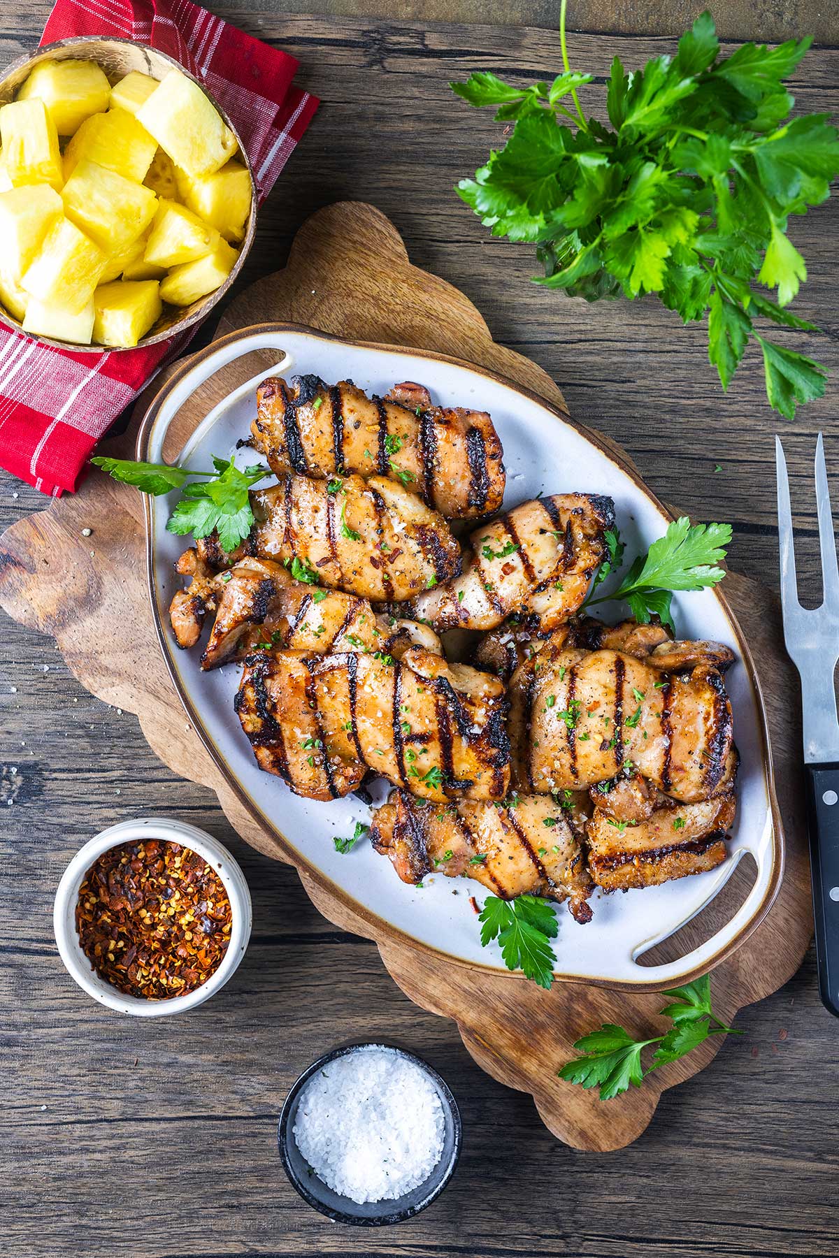 A platter of grilled chicken thighs garnished with parsley sits on a wooden board. Surrounding it are a bowl of pineapple chunks, a bowl of red pepper flakes, a bowl of salt, a fork, and fresh parsley.