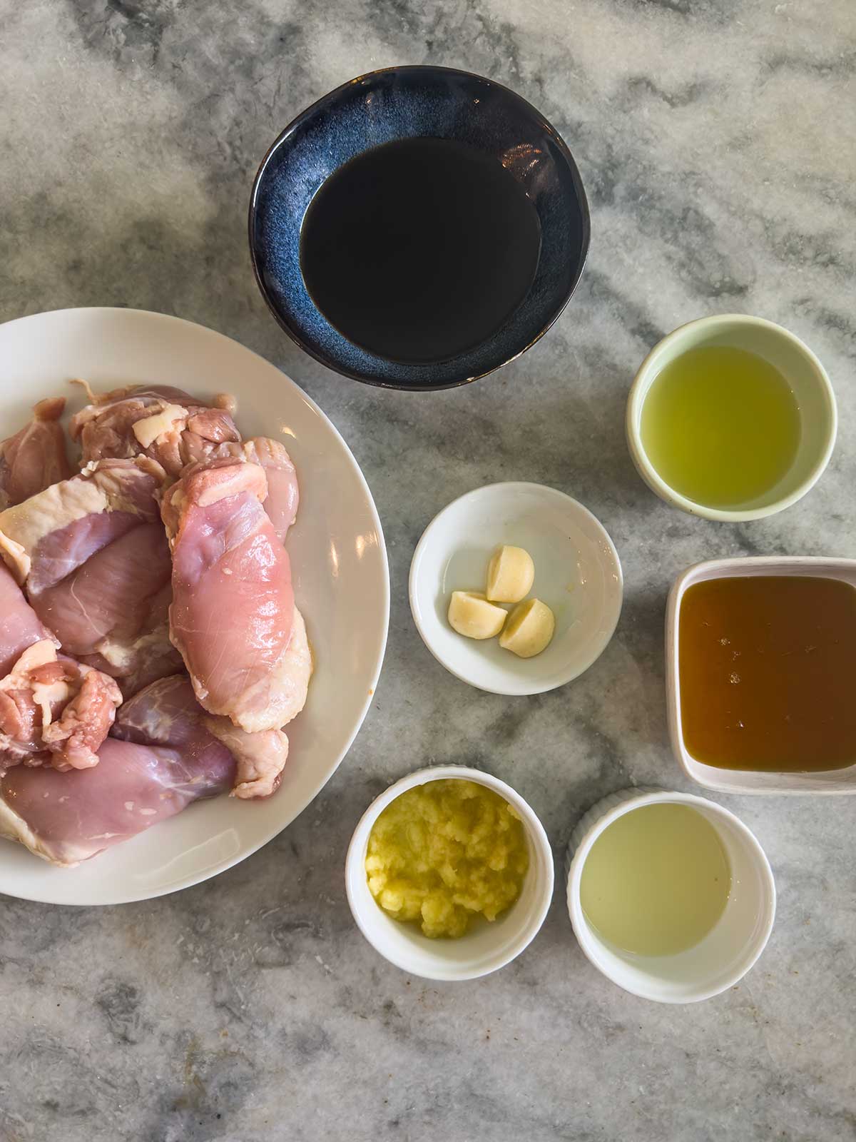 A top-down view of raw chicken thighs on a plate surrounded by small bowls containing soy sauce, olive oil, whole garlic cloves, honey, minced ginger, and vinegar on a marble surface.