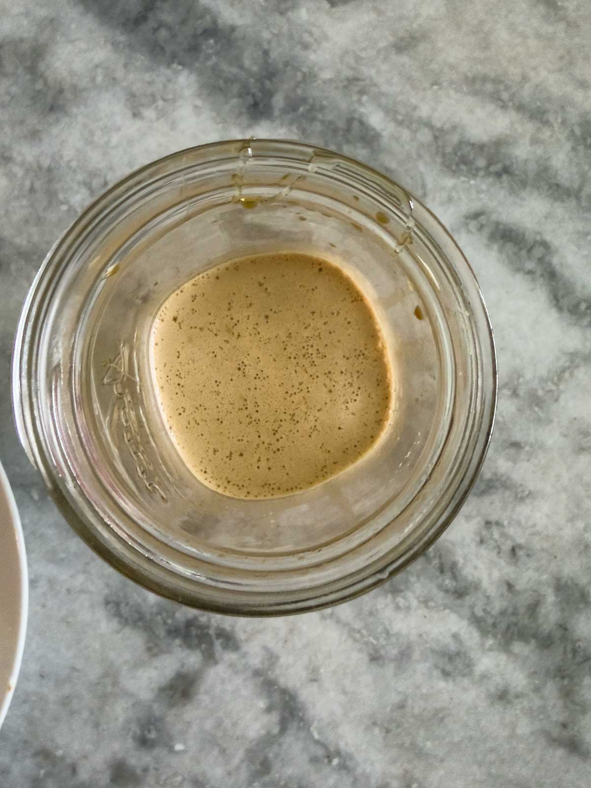 A glass jar with a small amount of creamy, light brown liquid at the bottom, viewed from above, sitting on a gray marble surface.