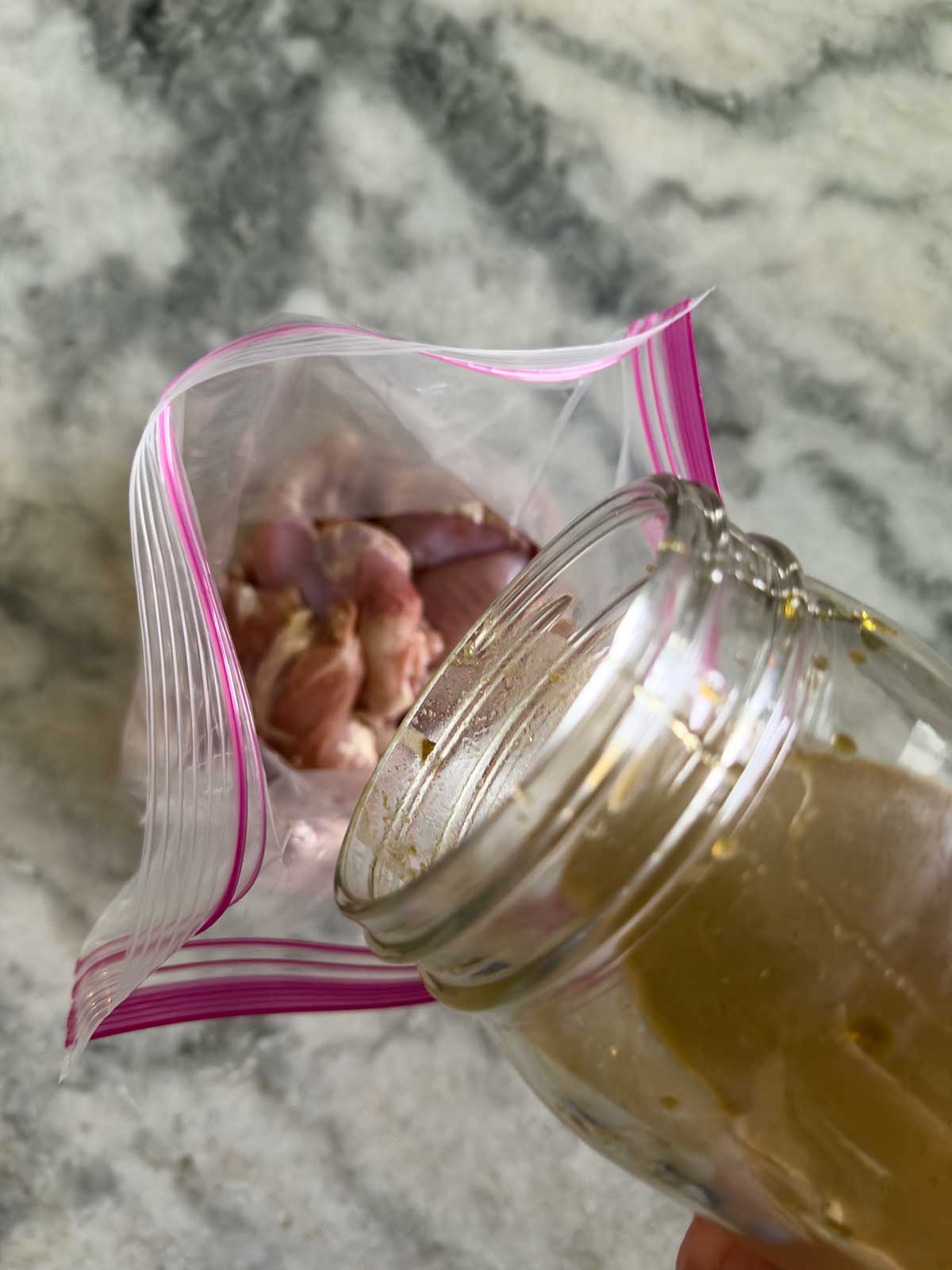 A hand pours marinade from a glass jar into a plastic zip-top bag containing raw chicken pieces, on a gray marble surface.