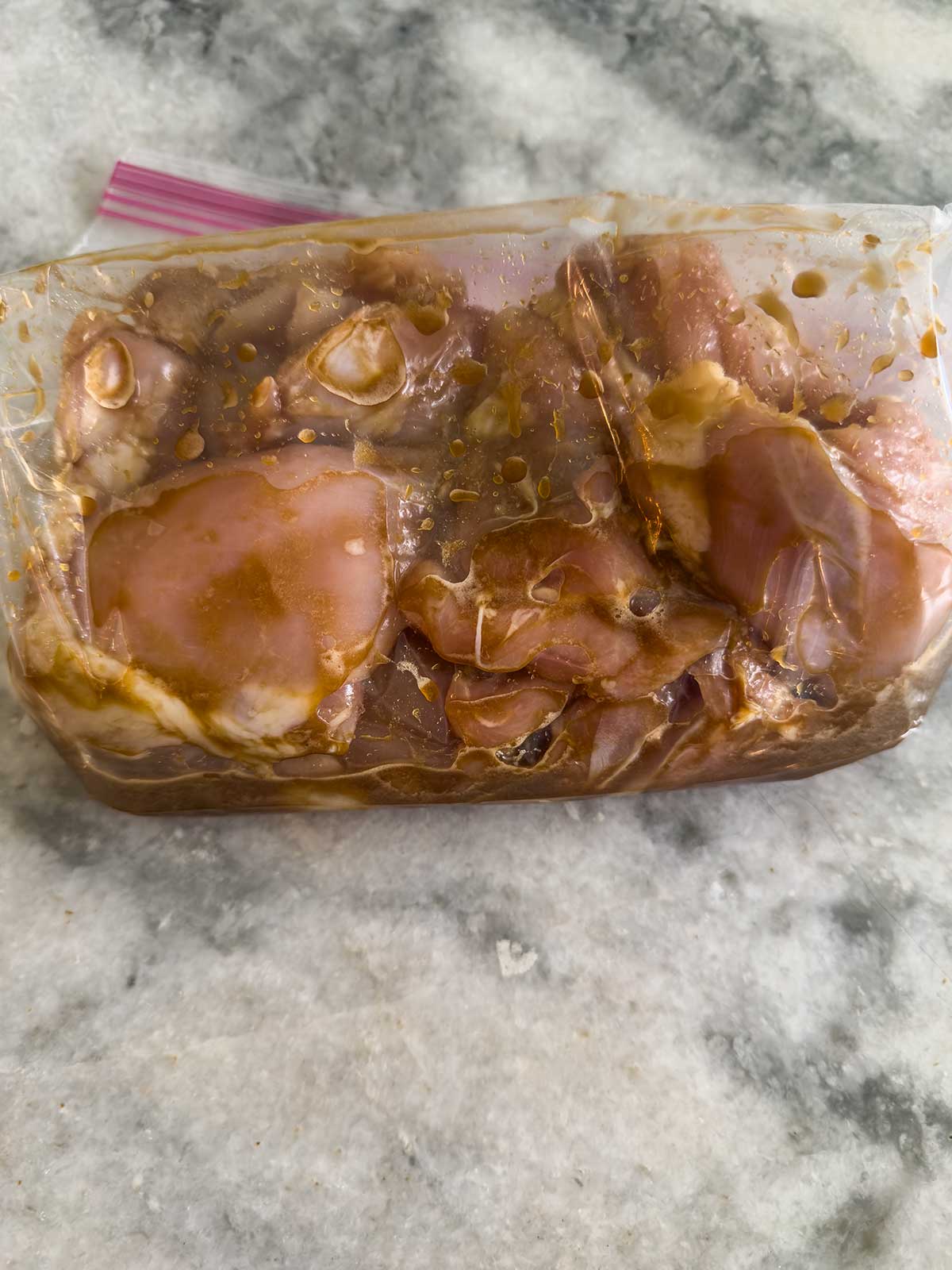 Raw chicken pieces marinating in a transparent ziplock bag with a brown sauce, placed on a gray marble countertop.