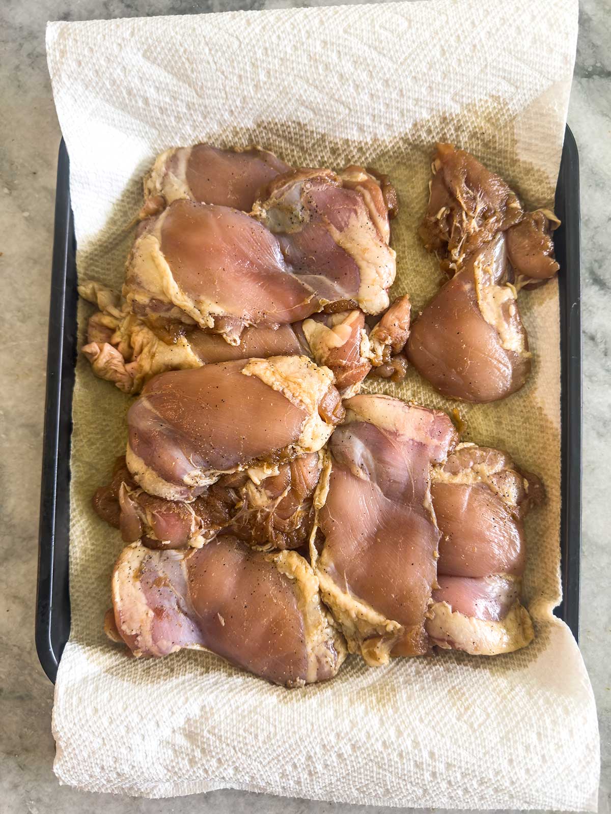 Raw marinated chicken thighs arranged on a baking tray lined with paper towels, ready for cooking. The tray is placed on a gray countertop.