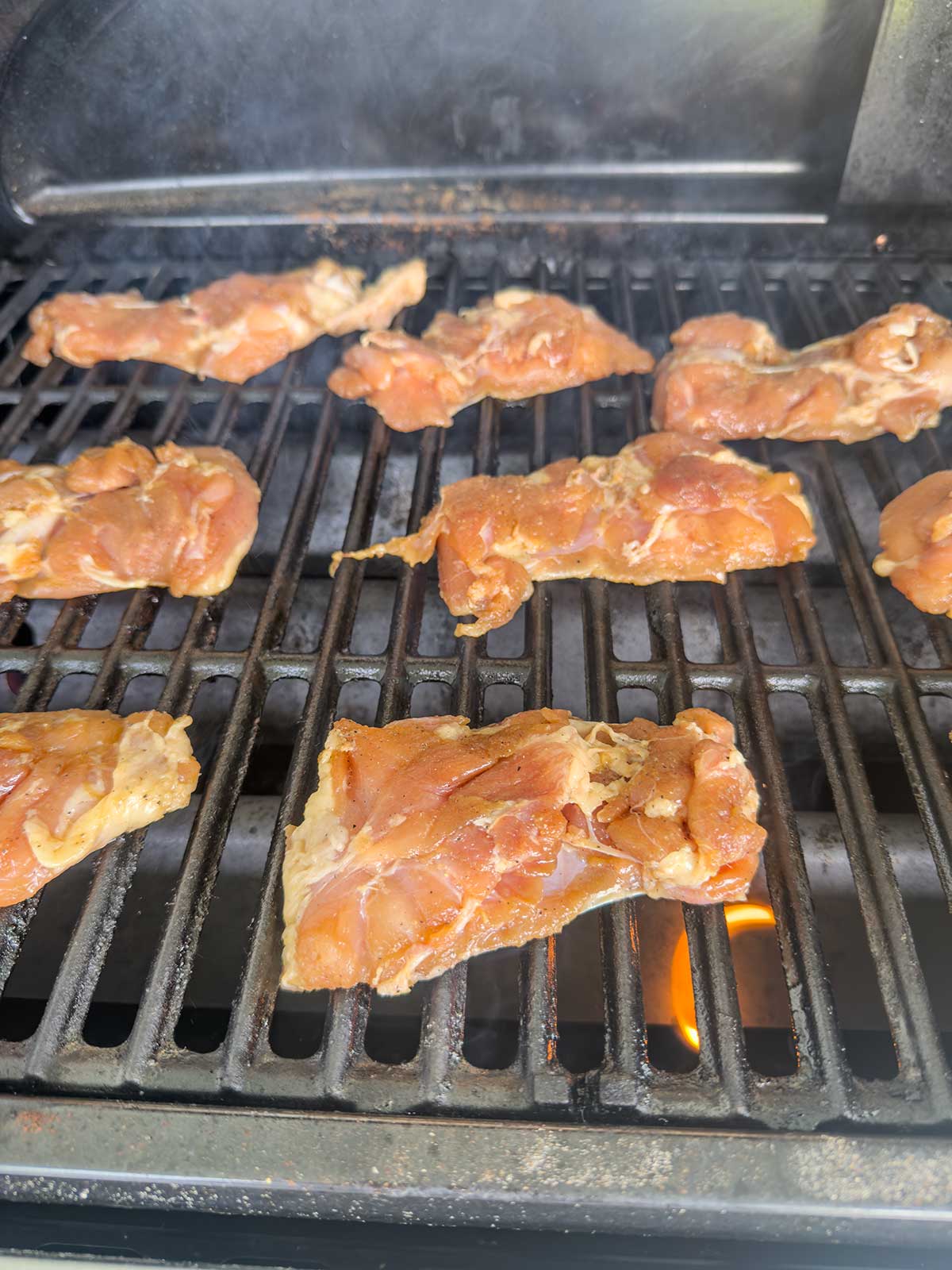 Several pieces of raw marinated chicken are being grilled on a barbecue, with flames visible under the grates. Smoke is rising, suggesting the grill is hot and cooking is in progress.