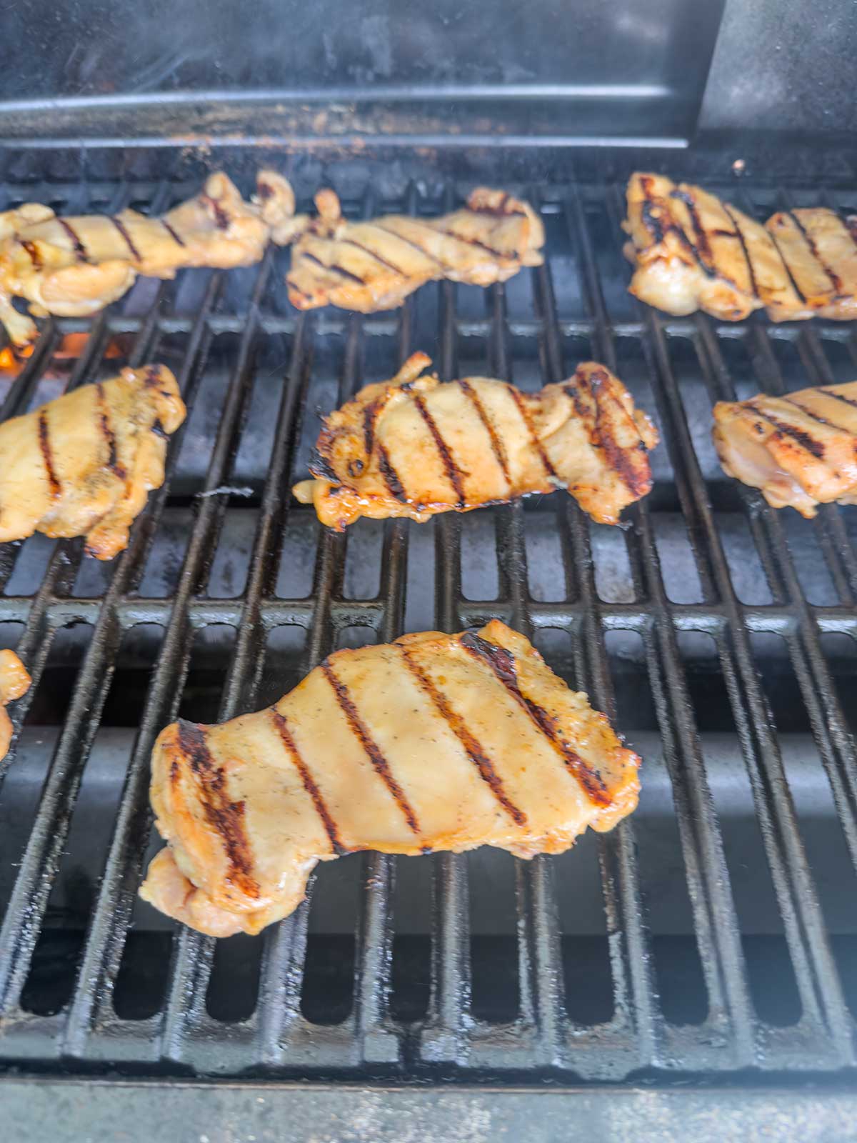 Several pieces of grilled chicken with dark grill marks are cooking on an outdoor barbecue grill. The chicken is arranged across the grates, some pieces more cooked than others.
