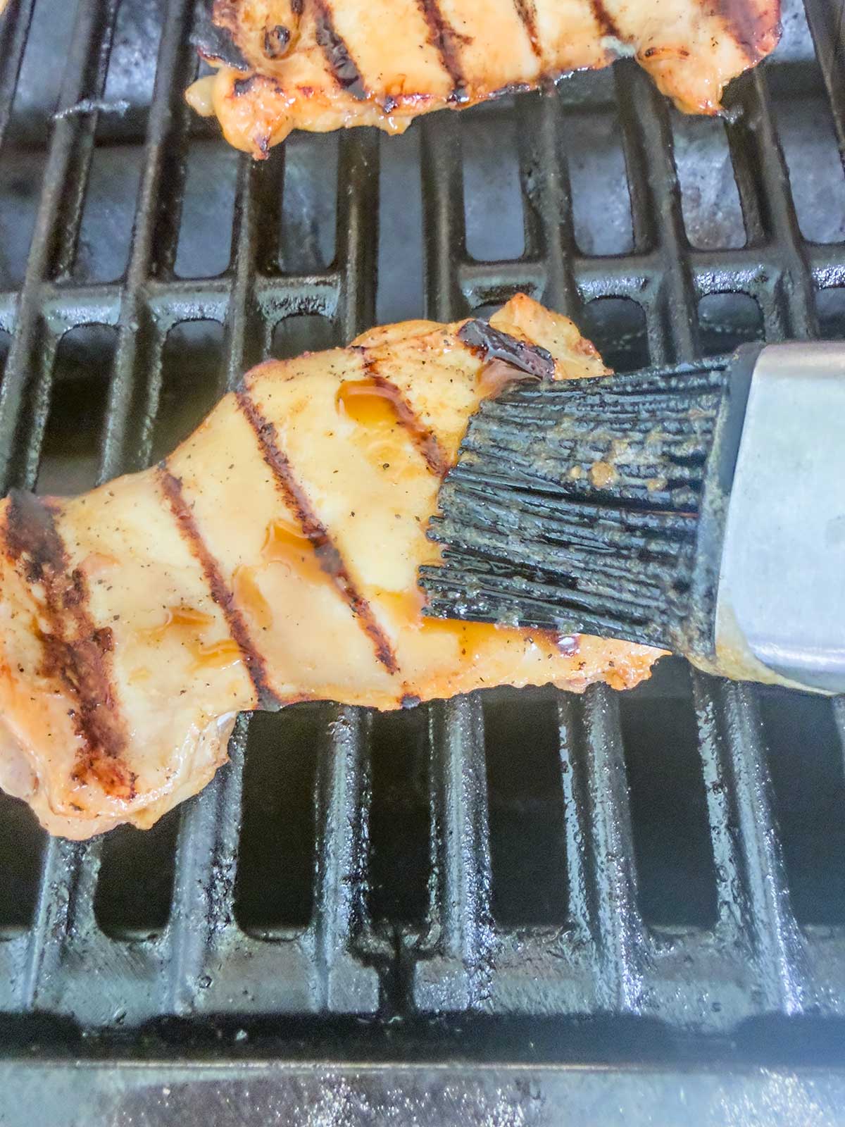 A piece of grilled chicken breast with char marks is being brushed with marinade or sauce while cooking on a grill.