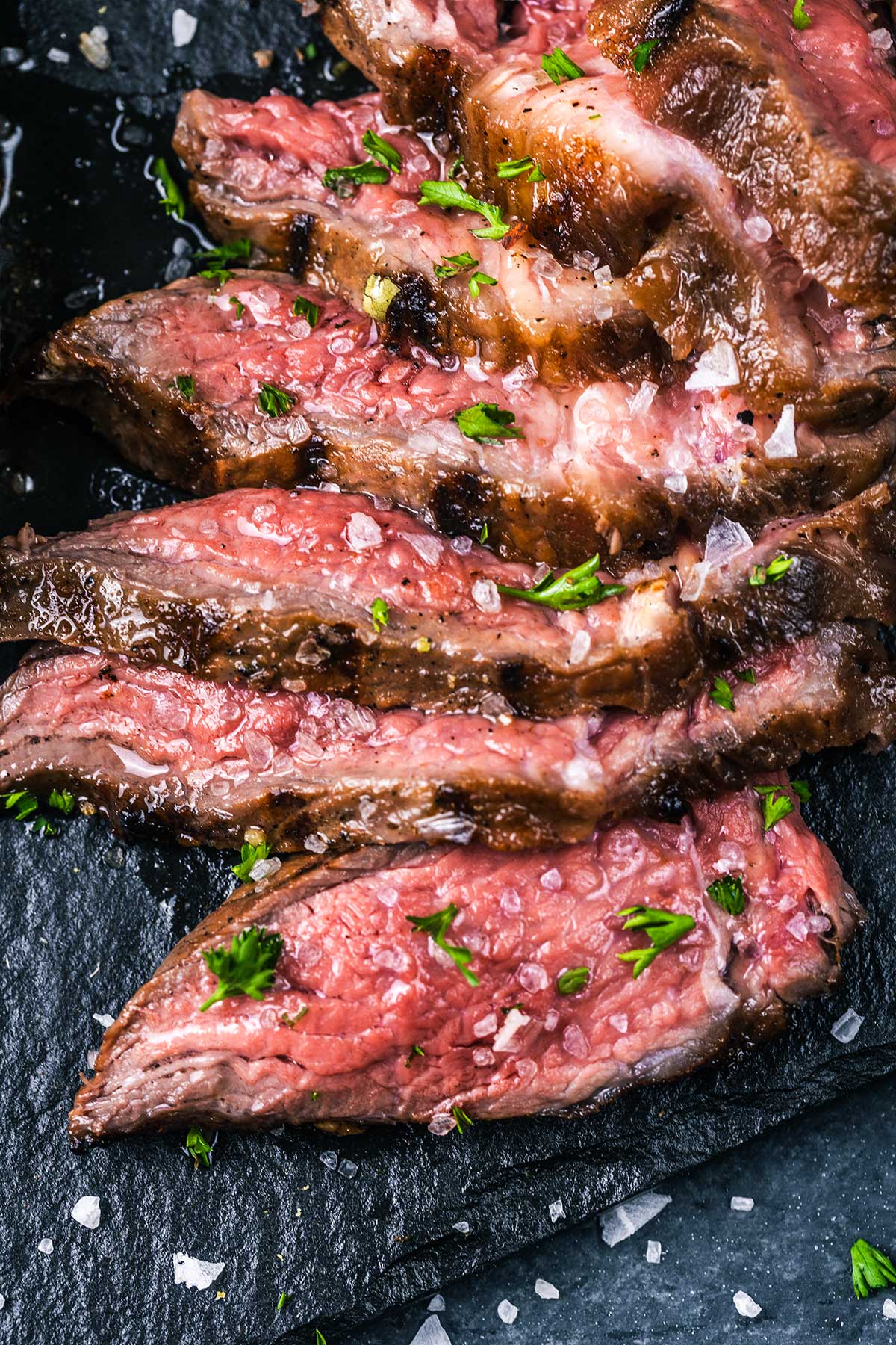 Sliced medium-rare grilled sirloin steak topped with coarse sea salt and chopped fresh herbs, served on a dark slate surface. The meat is juicy and pink inside with a charred outer crust.