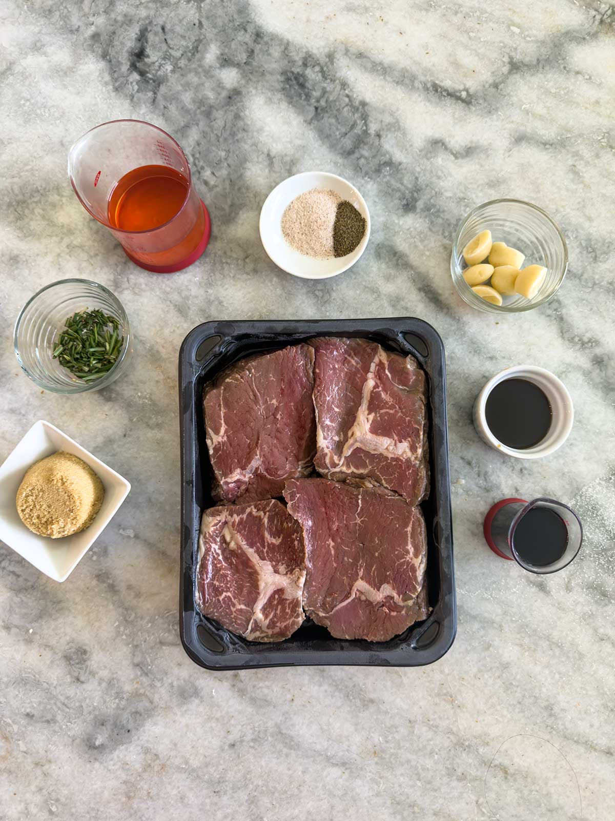 A tray of four raw beef chuck steaks, perfect alongside grilled sirloin steak, is surrounded by small bowls and cups filled with brown sugar, soy sauce, garlic cloves, herbs, spices, and liquids on a marble countertop.