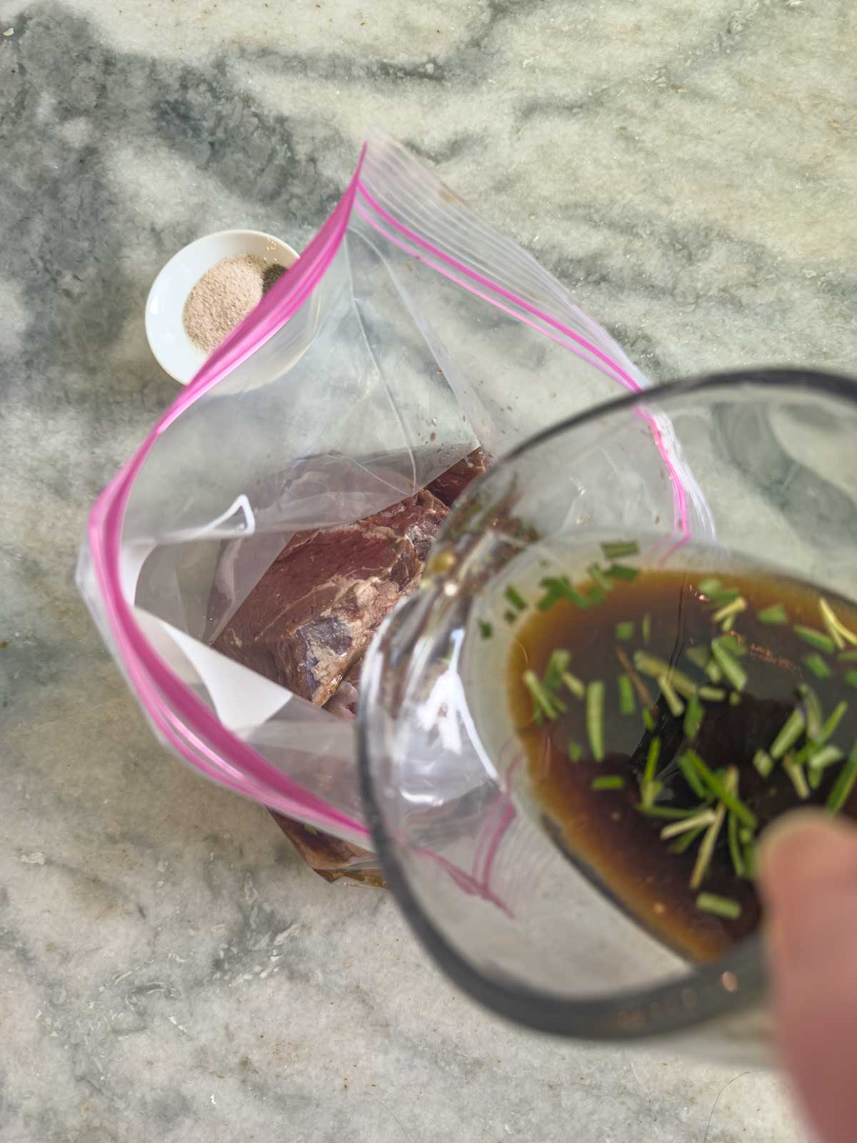 A hand pours a brown marinade with herbs into a plastic zip-top bag containing raw meat for grilled sirloin steak, with a small dish of salt nearby on a marble countertop.