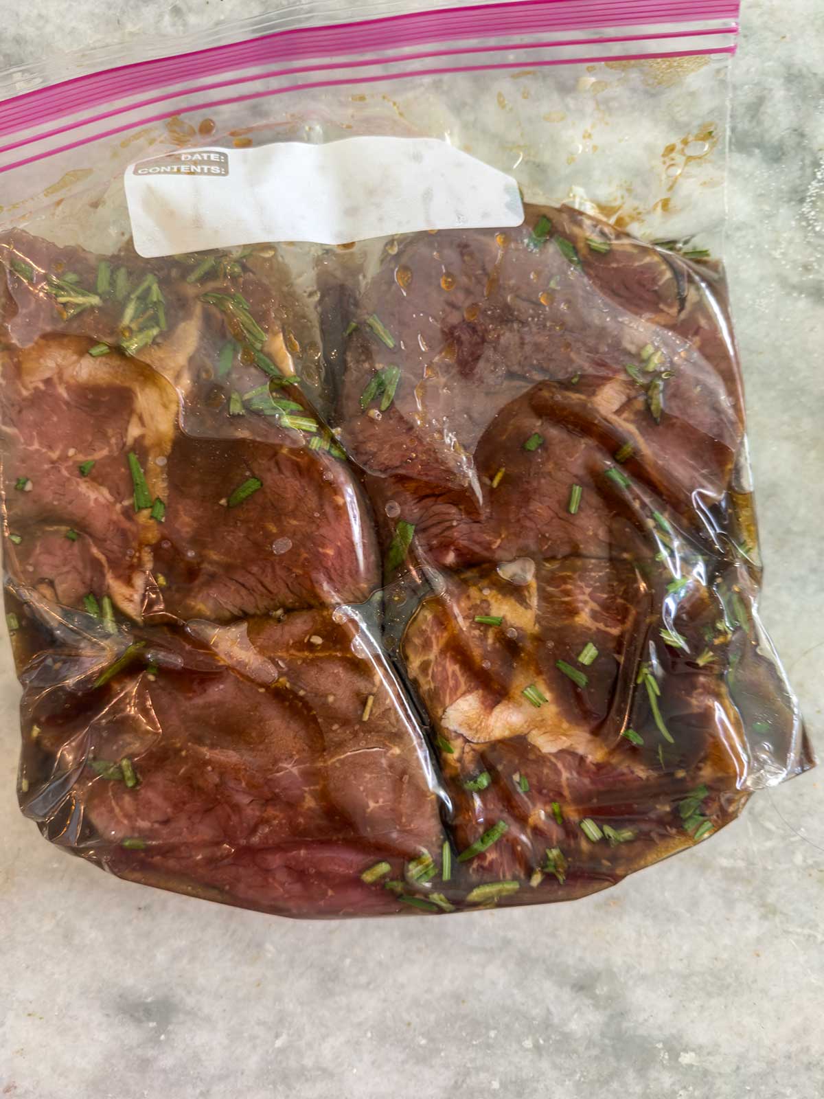 Raw meat marinating in a resealable plastic bag with herbs and marinade, perfect for preparing a delicious grilled sirloin steak, placed on a light-colored countertop.