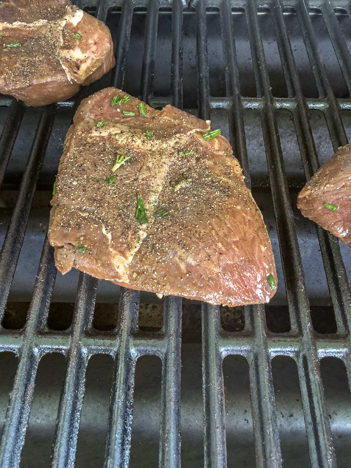 A seasoned grilled sirloin steak with herbs is sizzling on a barbecue grate. The uncooked meat rests diagonally on the metal bars, with other pieces of steak partially visible nearby.