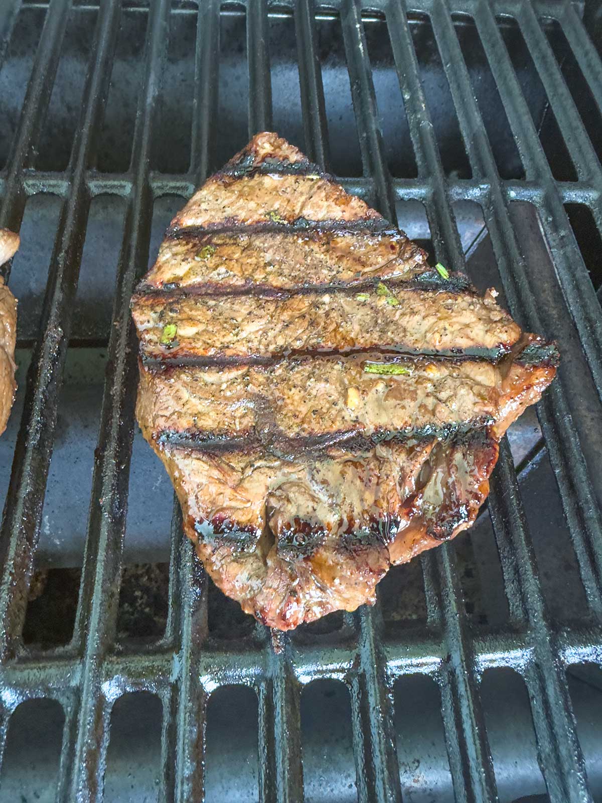 A grilled sirloin steak with visible char marks is cooking on a metal barbecue grill, seasoned with herbs and spices.