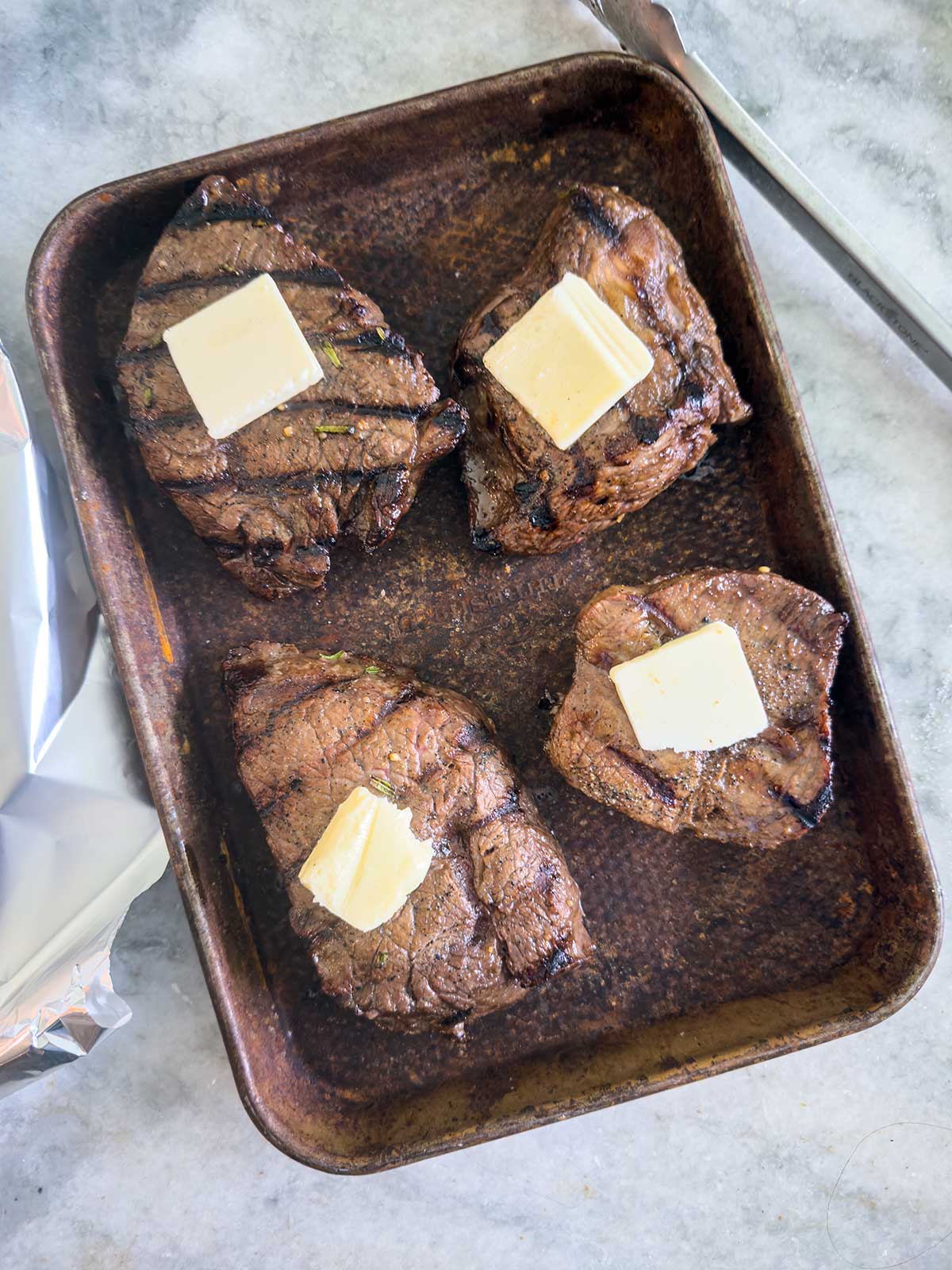 Four grilled sirloin steaks in a baking pan, each topped with a square of melting butter, rest on a light-colored countertop with foil and a knife visible nearby.