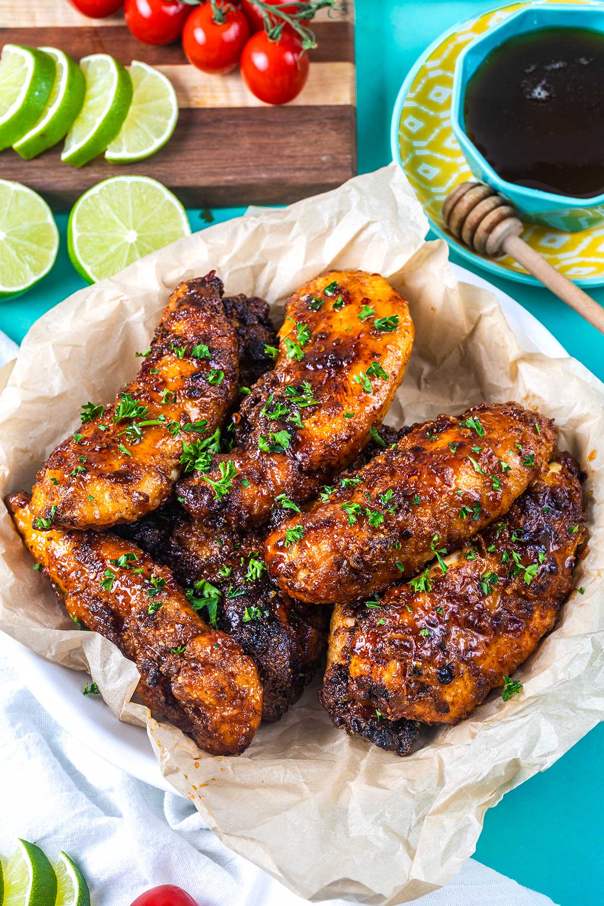 A plate of grilled glazed chicken breasts garnished with chopped parsley, placed on parchment paper. Sliced limes, cherry tomatoes, and a bowl of dark sauce with a honey dipper are nearby on a vibrant blue surface.