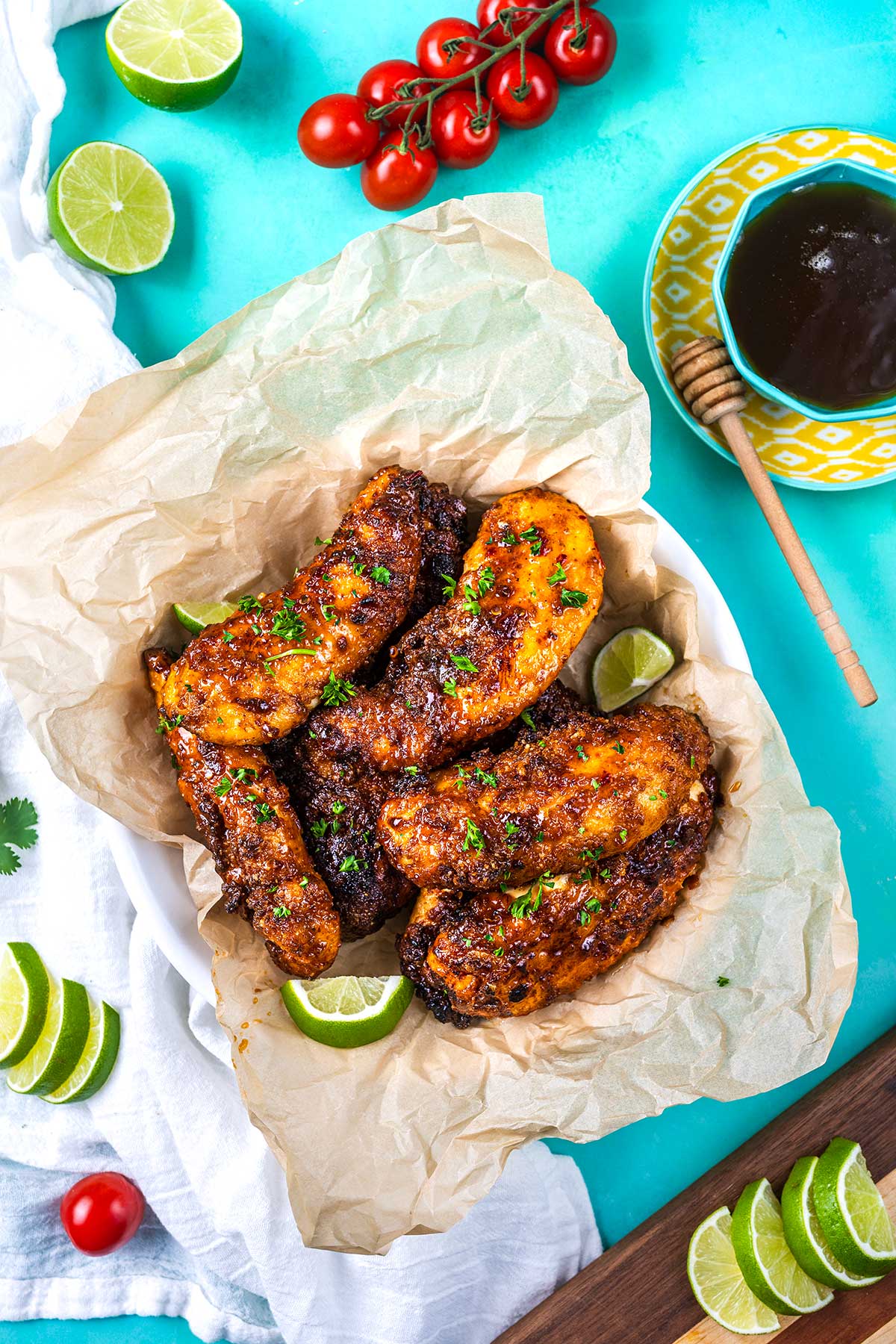 A bowl lined with parchment paper holds crispy glazed chicken tenders, garnished with herbs and lime wedges. Around it are lime slices, cherry tomatoes on the vine, a bowl of dark dipping sauce, and a honey dipper.
