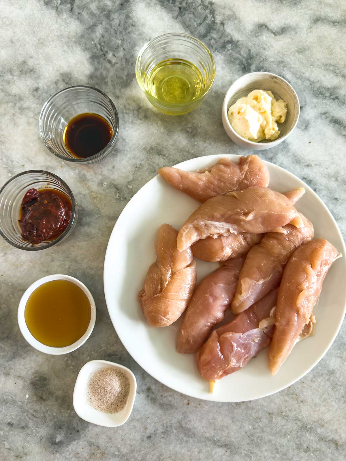 Raw chicken tenders on a white plate surrounded by small bowls of oil, mayonnaise, chipotle peppers in sauce, soy sauce, honey, and a dish of seasoning, all on a marble countertop.