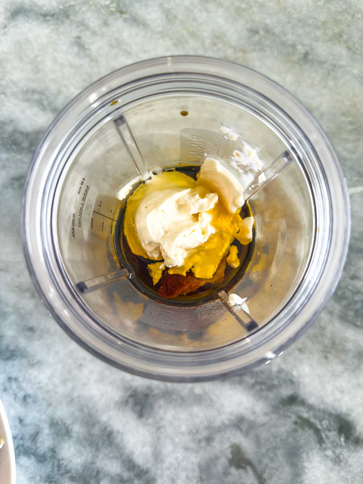 A top-down view of a blender cup containing mayonnaise, mustard, honey, and a dark liquid, placed on a gray marble countertop.