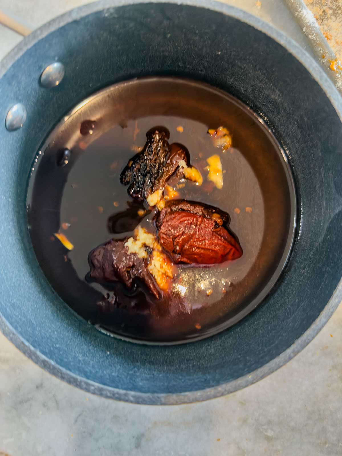 A black cooking pot contains a dark liquid with several chunks of what appears to be burnt and partially cooked food, sitting on a gray countertop.