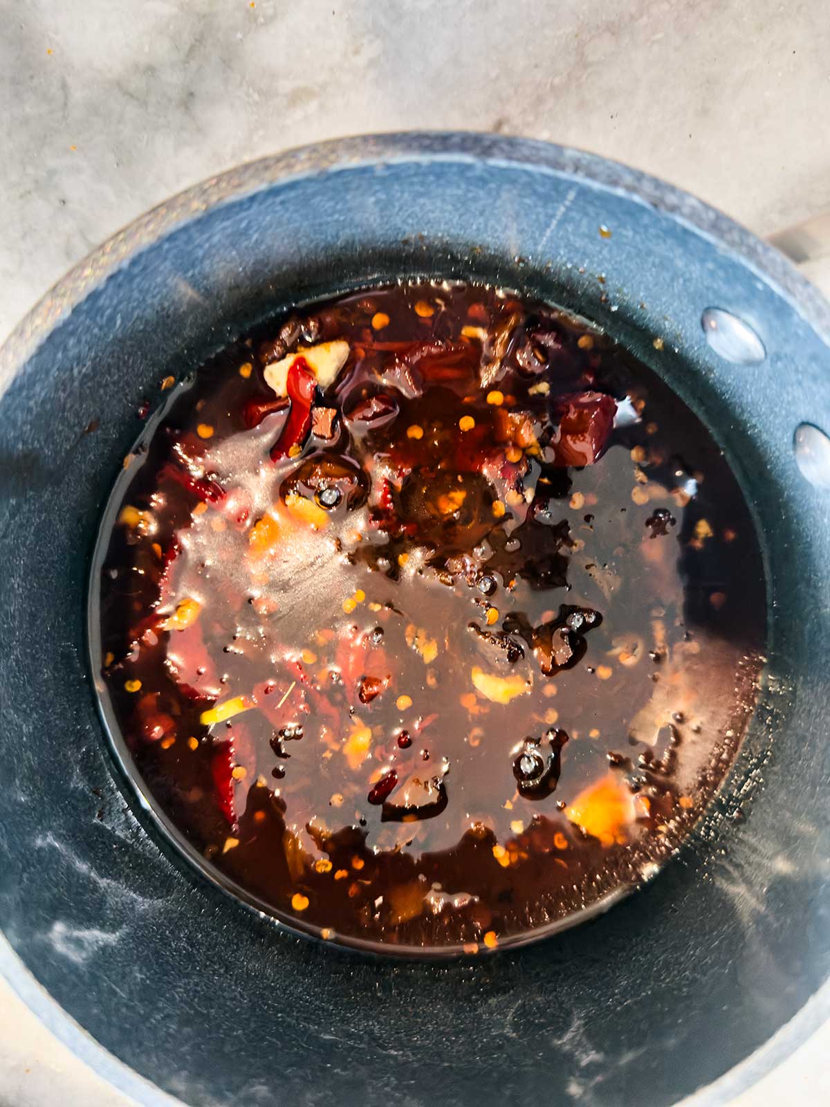 A dark, glossy sauce with chili flakes, garlic, and other seasonings simmers in a dark pot.