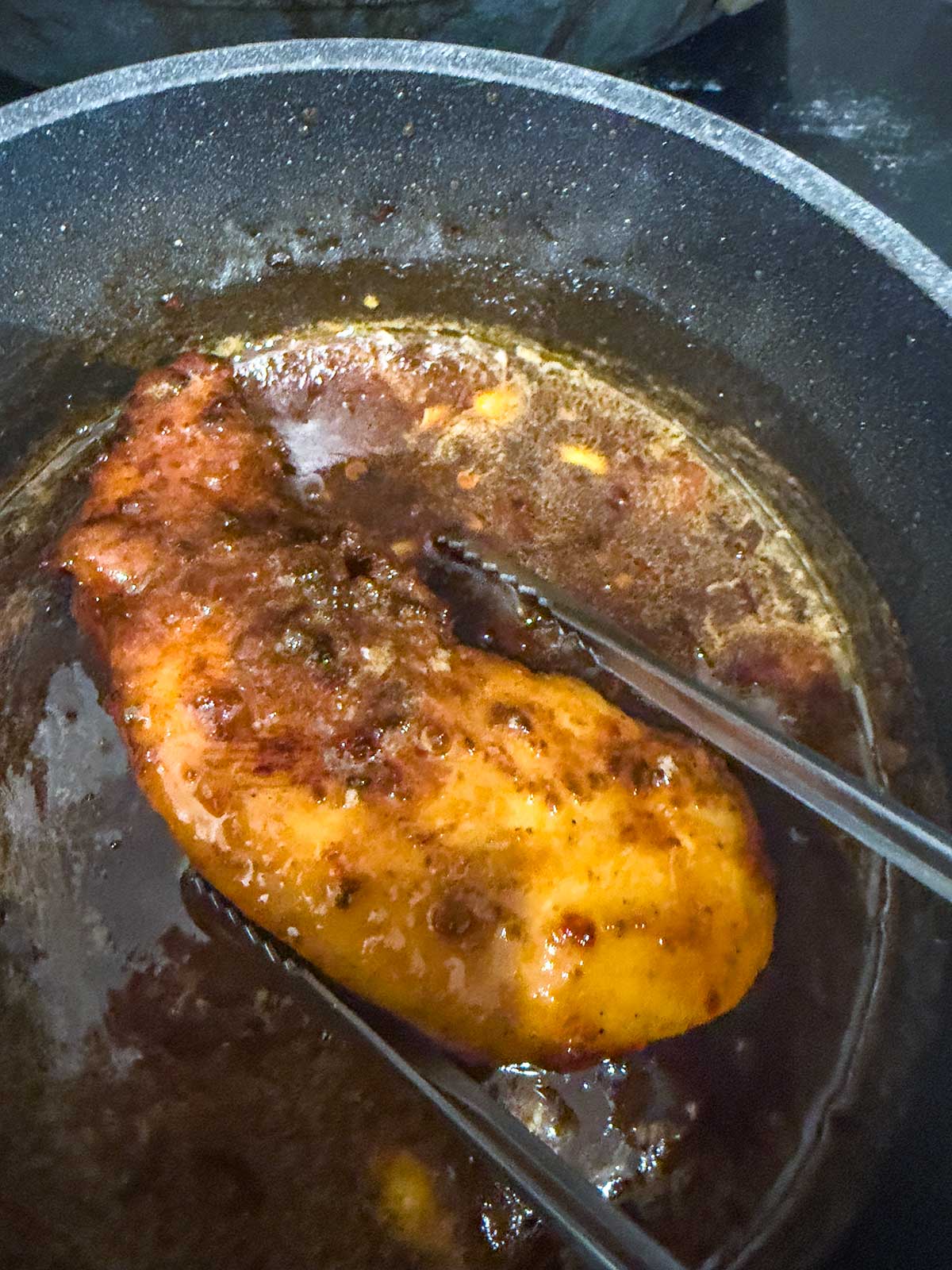 A cooked chicken breast is held with metal tongs over a pan filled with a dark, bubbling sauce. The chicken is coated in the sauce and appears tender and seasoned.