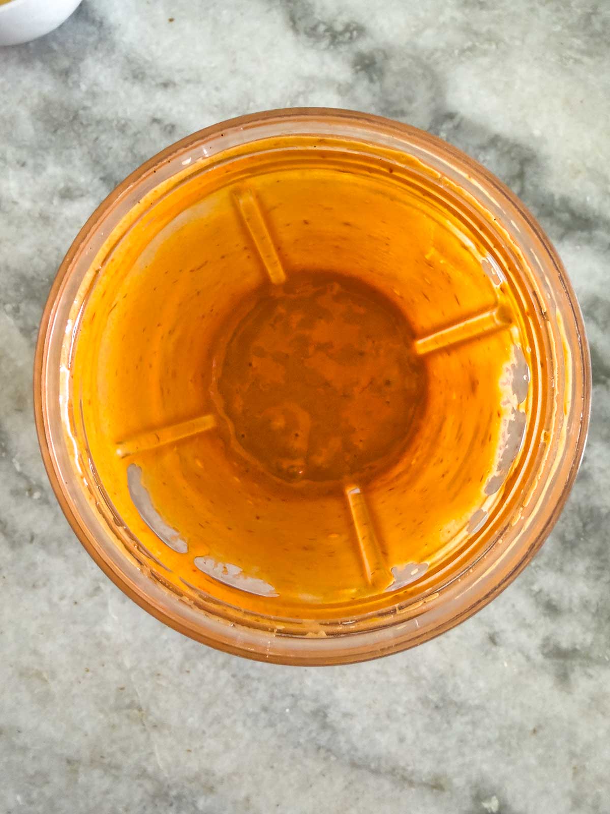 Top view of a blender cup containing a small amount of orange-colored sauce or dressing, with some residue coating the sides. The blender is placed on a light gray marble countertop.
