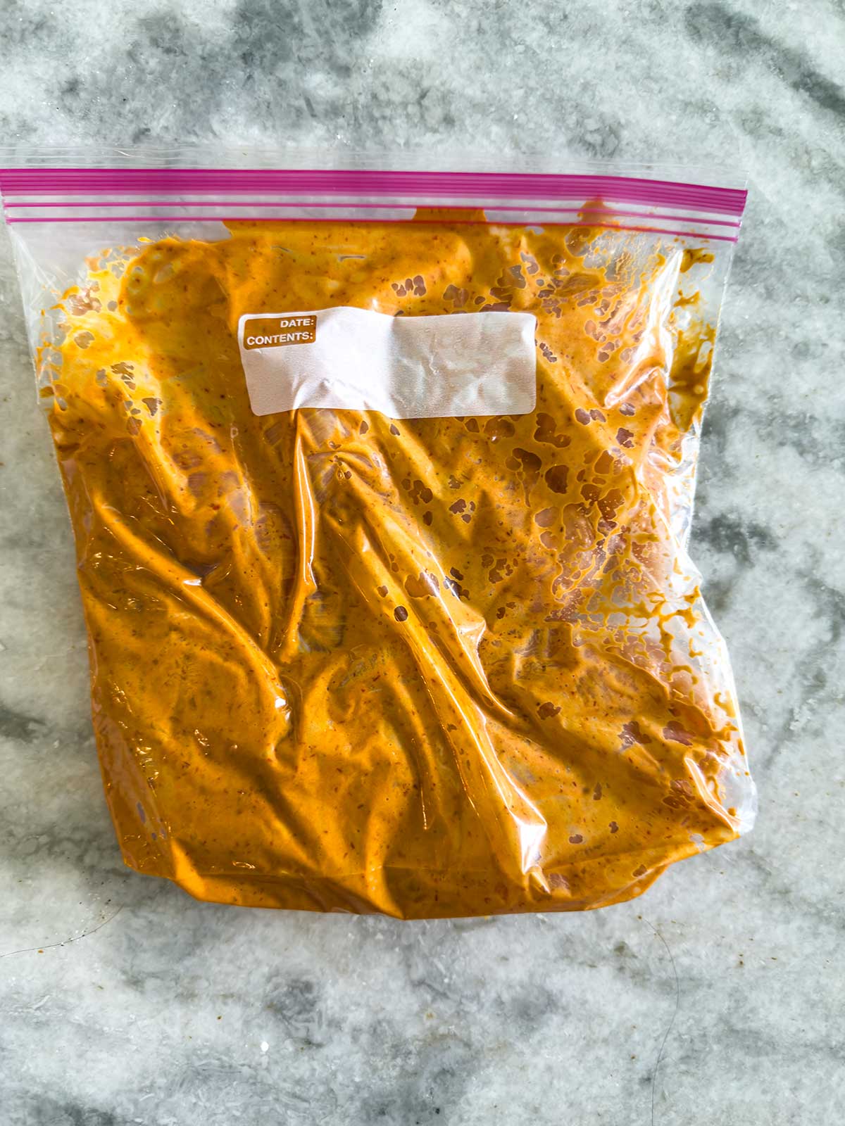 A resealable plastic bag filled with a thick, orange-yellow marinade sits on a gray marble surface. A white label is attached to the bag for writing the date and contents.