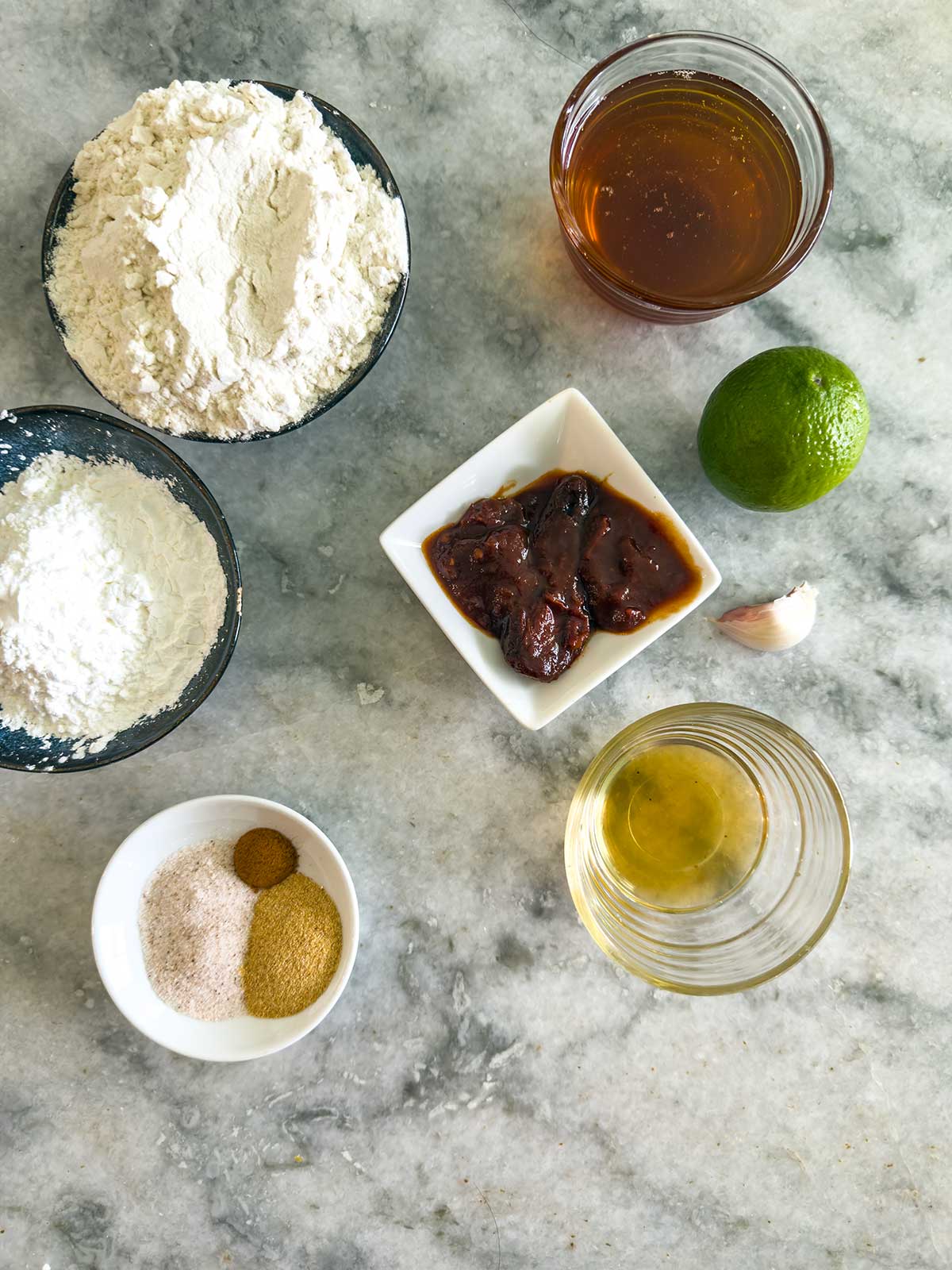 A marble countertop with bowls containing flour, cornstarch, spices, chipotle peppers, a glass of oil, a glass of brown liquid, a lime, and a garlic clove arranged neatly.