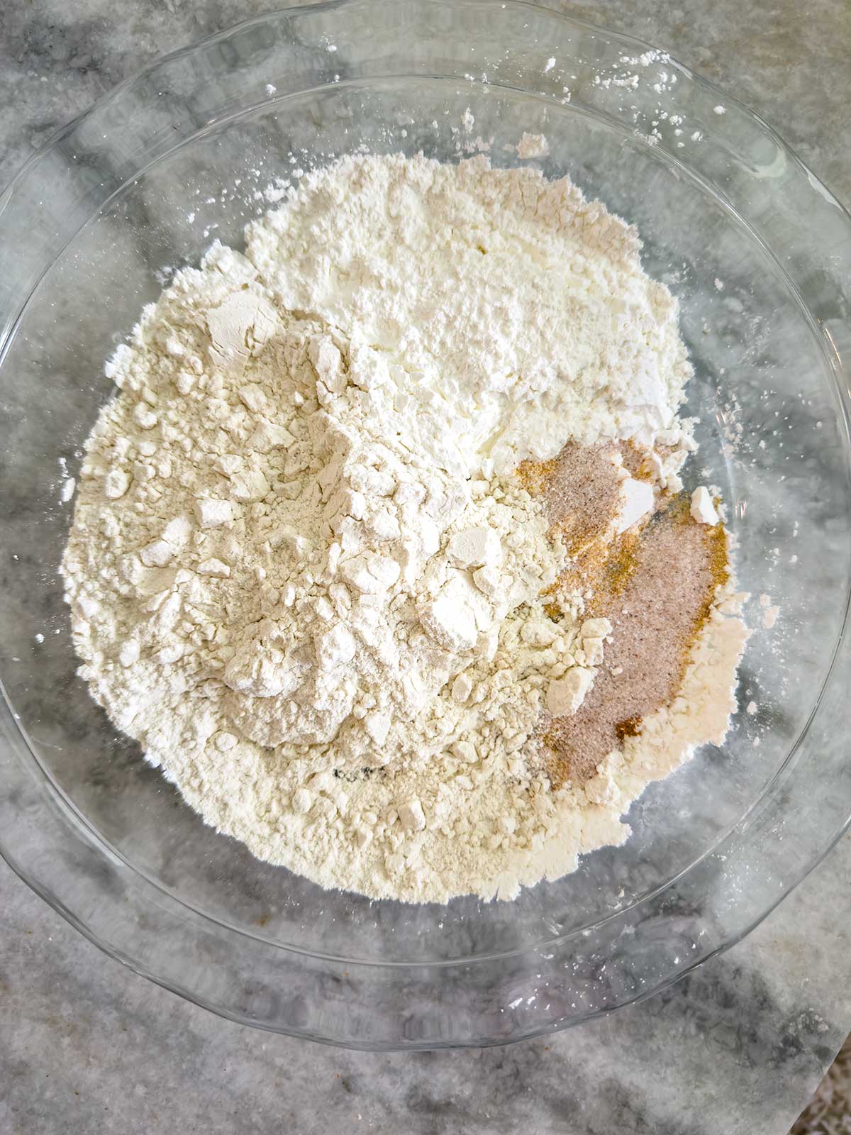 A pile of white flour sits in a clear glass bowl next to a small pile of brown sugar and spices.