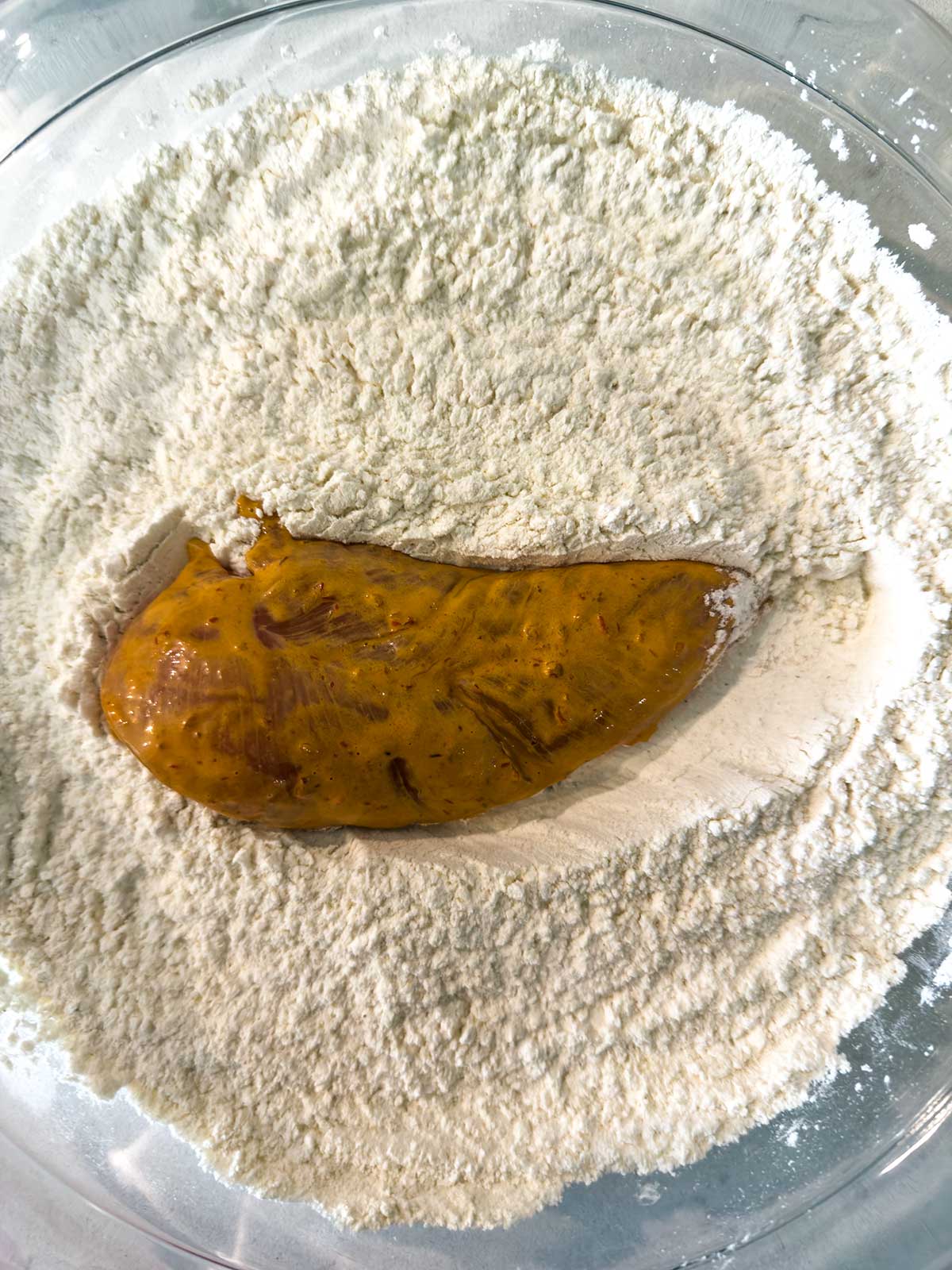 A marinated chicken breast is placed on a bed of flour in a glass mixing bowl, ready to be coated before cooking.