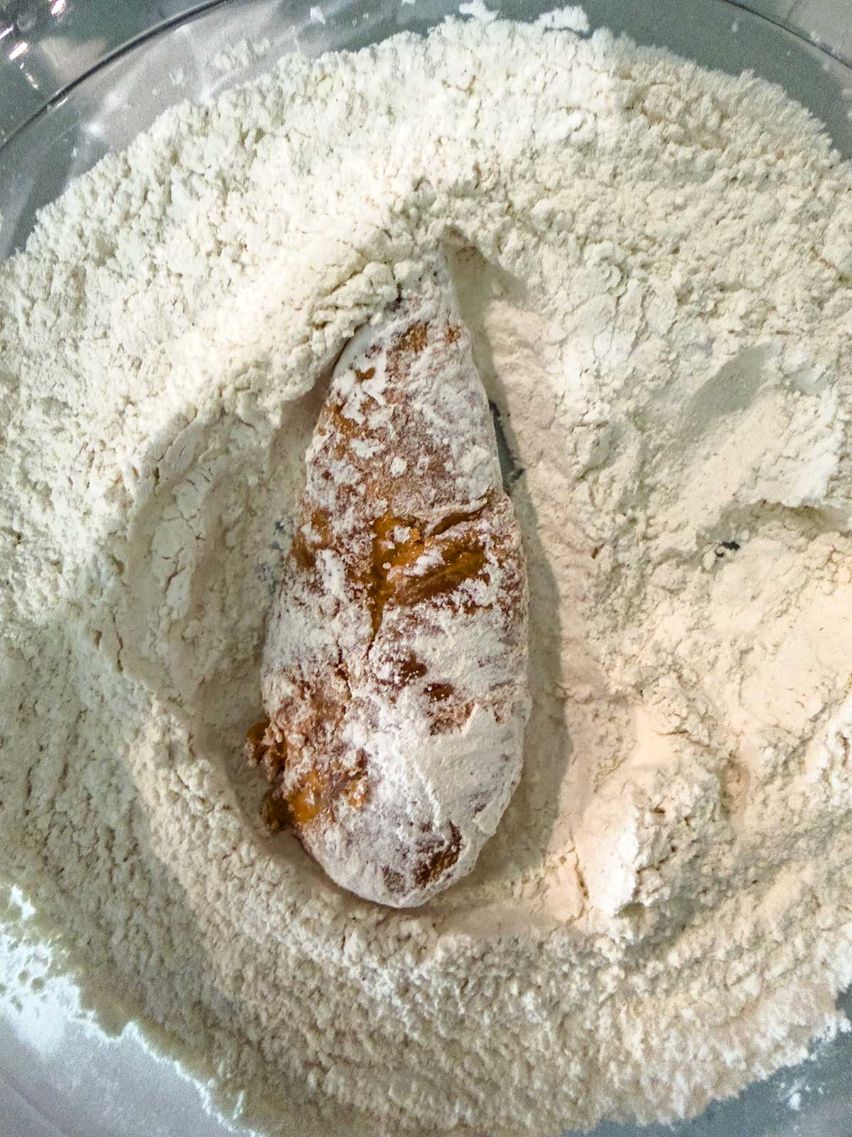 A piece of food, possibly chicken, coated in a sticky brown marinade, sits in a bowl of white flour, ready to be breaded.