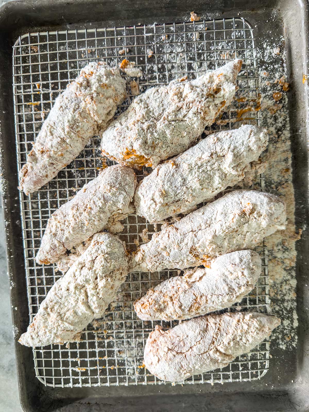 Eight uncooked chicken tenders coated in flour sit on a wire rack over a baking sheet, ready to be cooked.