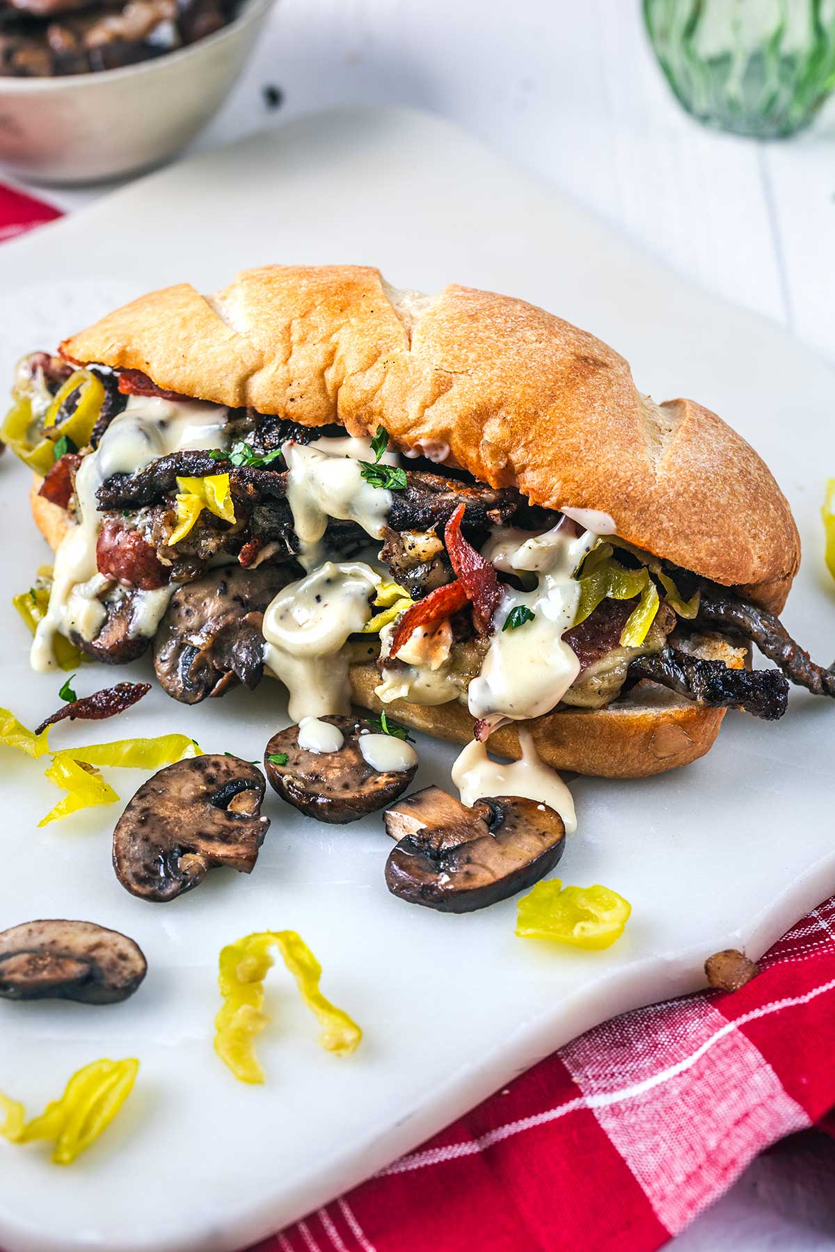 A toasted Italian cheesesteak sandwich filled with sautéed mushrooms, sliced peppers, pepperoni, and creamy sauce sits on a white cutting board, with mushrooms, pepper slices, and a red napkin nearby.