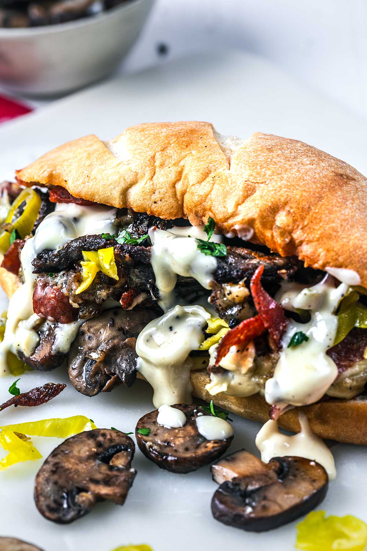 A toasted sandwich filled with sliced mushrooms, bacon, yellow peppers, and topped with creamy white sauce, sits on a white surface with extra mushroom slices and pepper rings beside it.