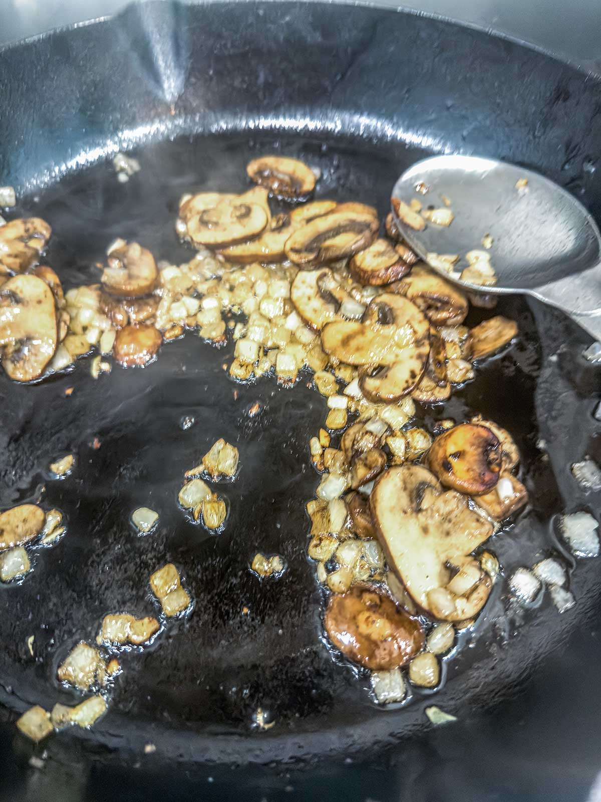 Sliced mushrooms and diced onions sautéing in a black cast iron skillet, with a metal spoon resting on the side of the pan.