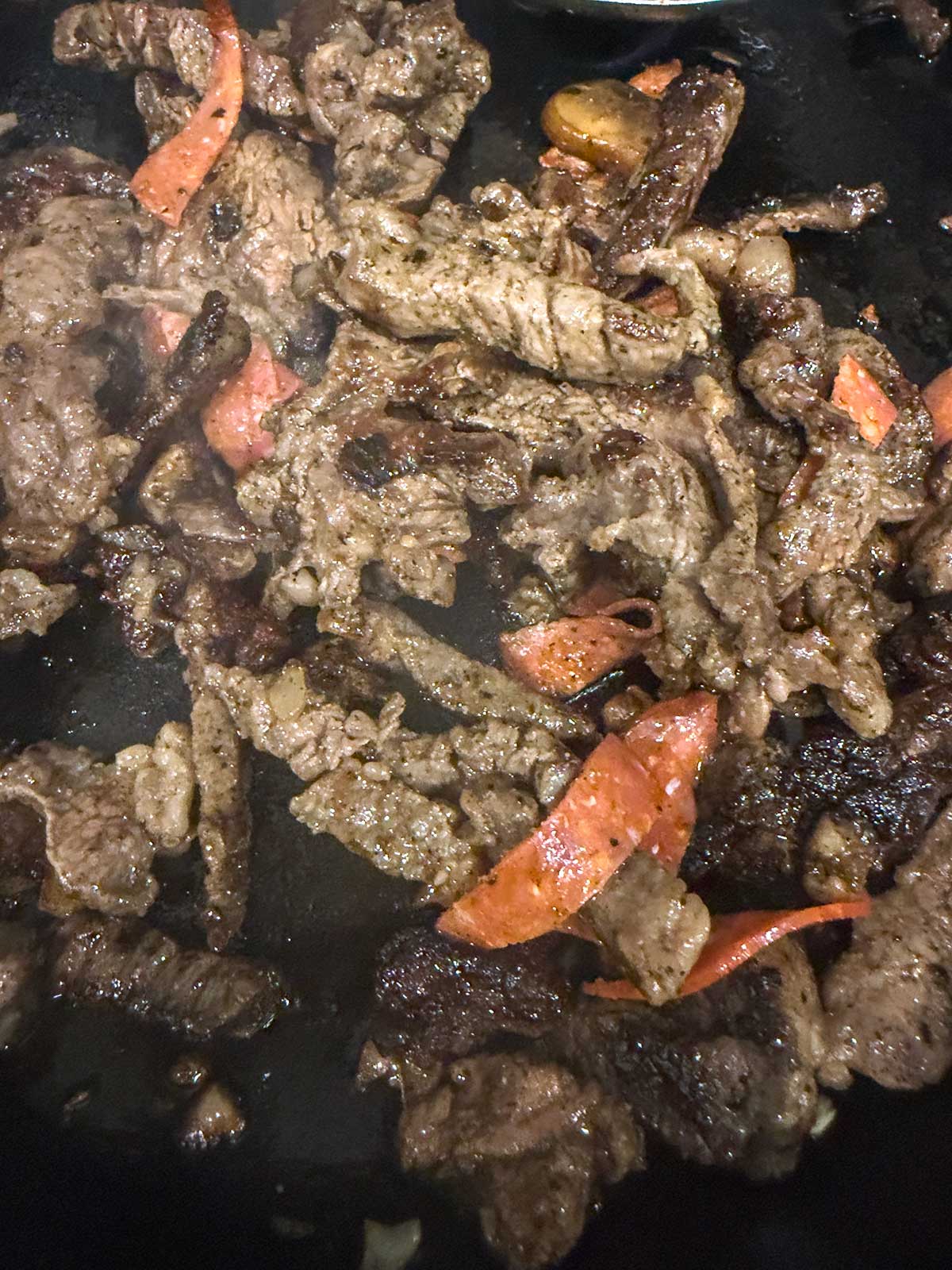 Sliced beef strips and pieces of red bell pepper cook together in a dark pan, seasoned and seared, with visible browning and a slightly oily texture.