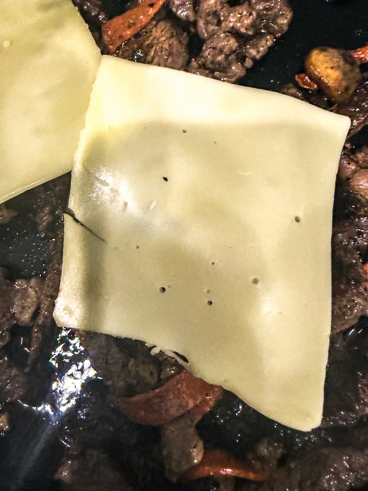 A close-up of sliced Swiss cheese melting over cooked beef strips and sautéed vegetables in a pan.
