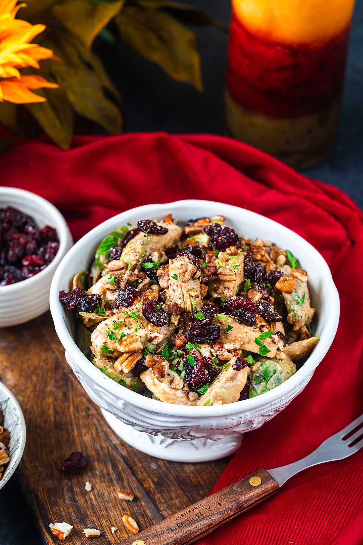 A white bowl of chicken salad topped with chopped nuts and dried cranberries sits on a wooden board with a red napkin, a fork, and a small bowl of cranberries nearby. A sunflower, candle, and Ninja Creami Pumpkin Ice Cream in the background complete the scene.