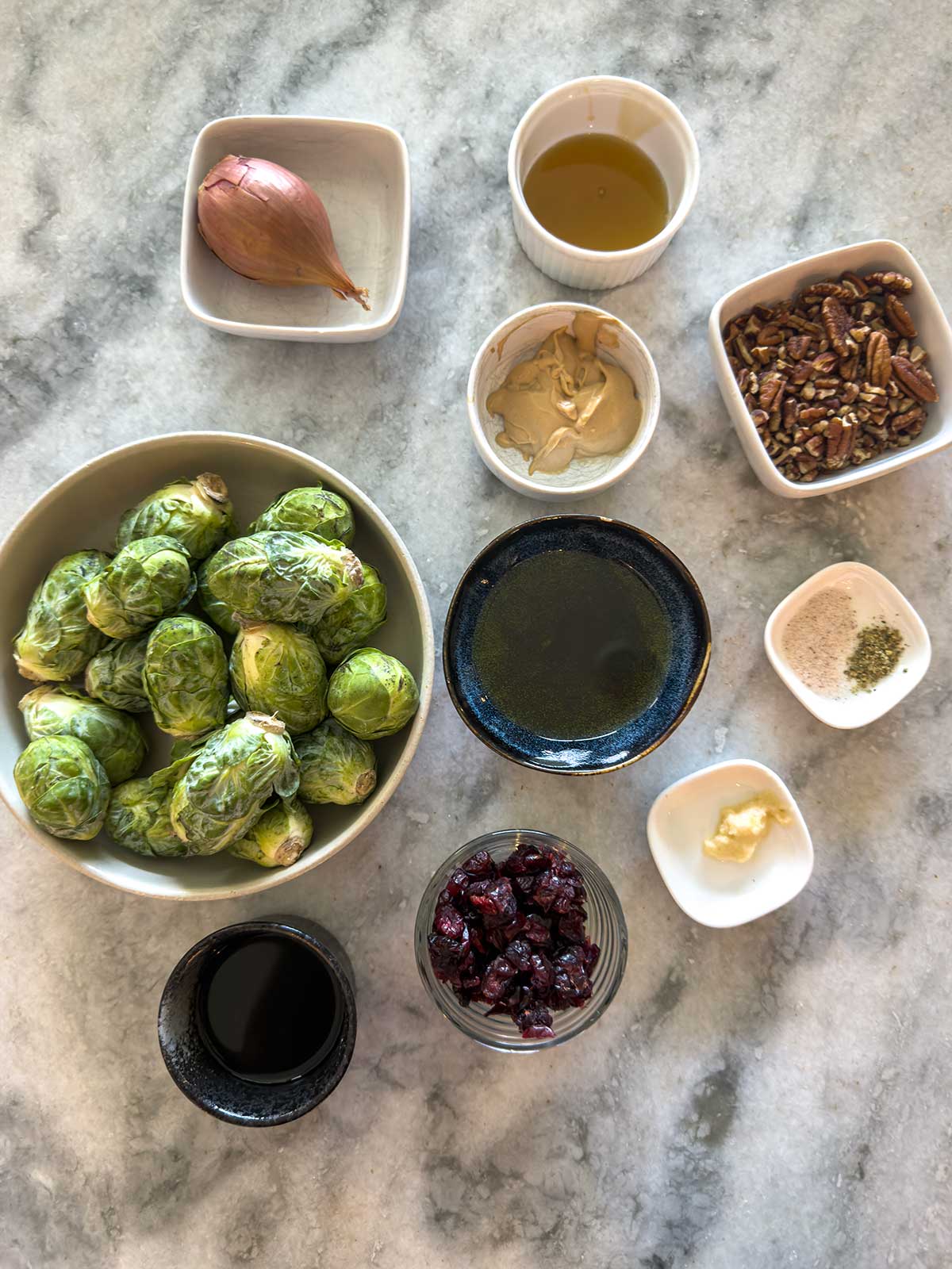Overhead view of ingredients on a marble surface, including Brussels sprouts, a shallot, olive oil, Dijon mustard, chopped pecans, dried cranberries, butter, balsamic vinegar, salt, and pepper.
