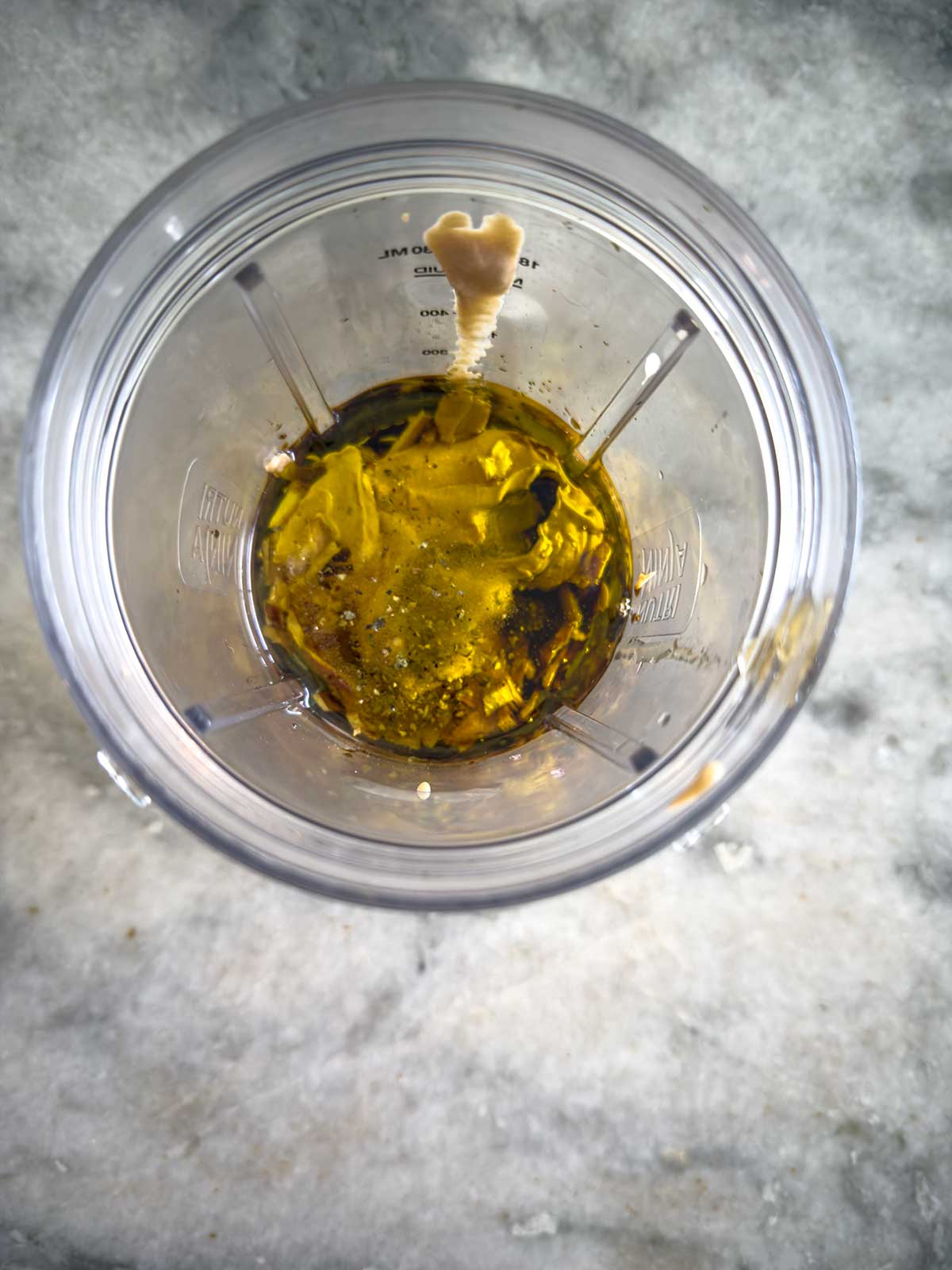 A clear blender cup with olive oil, mustard, and seasonings inside, viewed from above on a gray marble countertop.