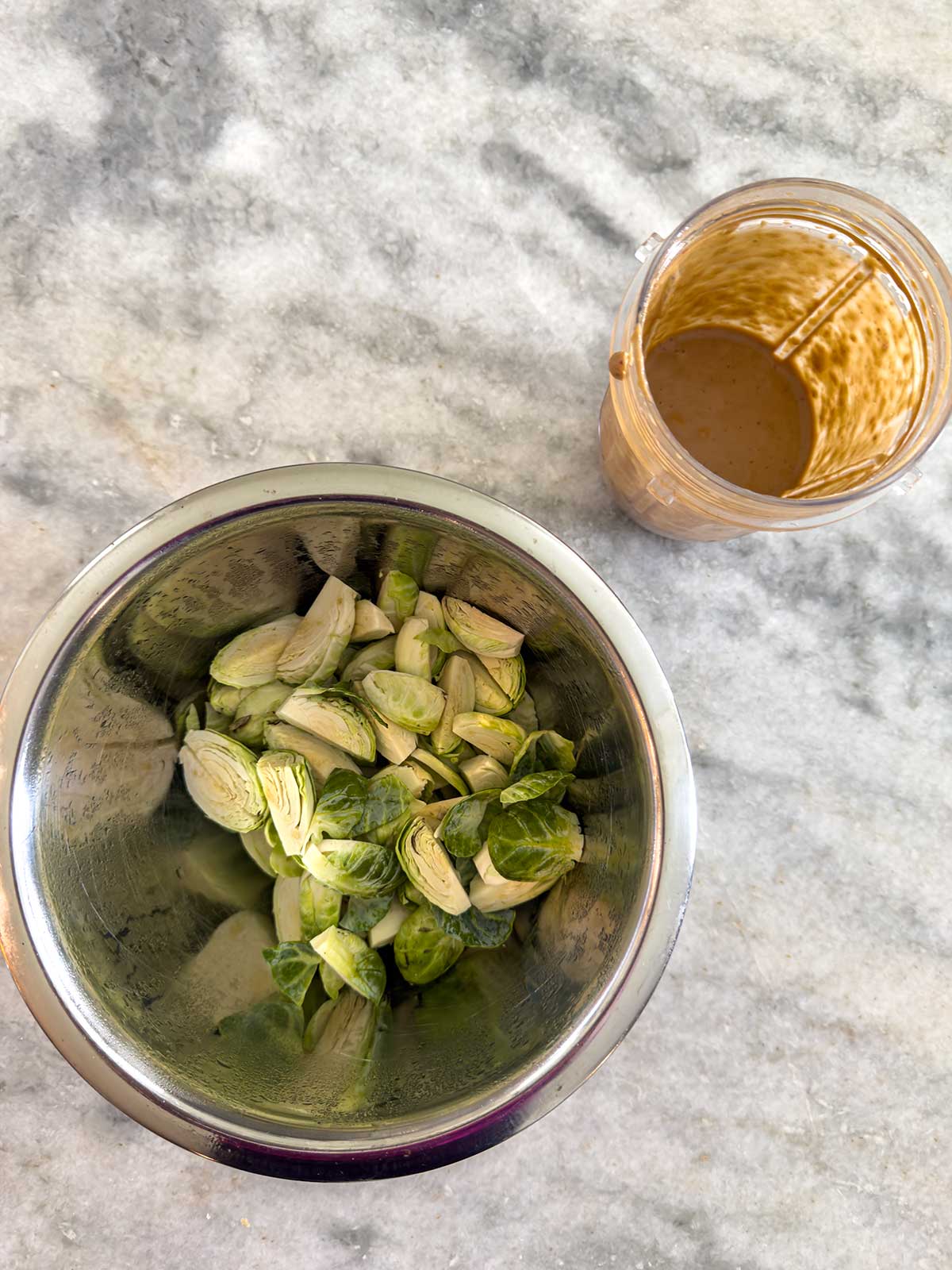 A metal bowl filled with halved Brussels sprouts sits on a marble counter next to a small blender cup containing a light brown sauce or dressing.