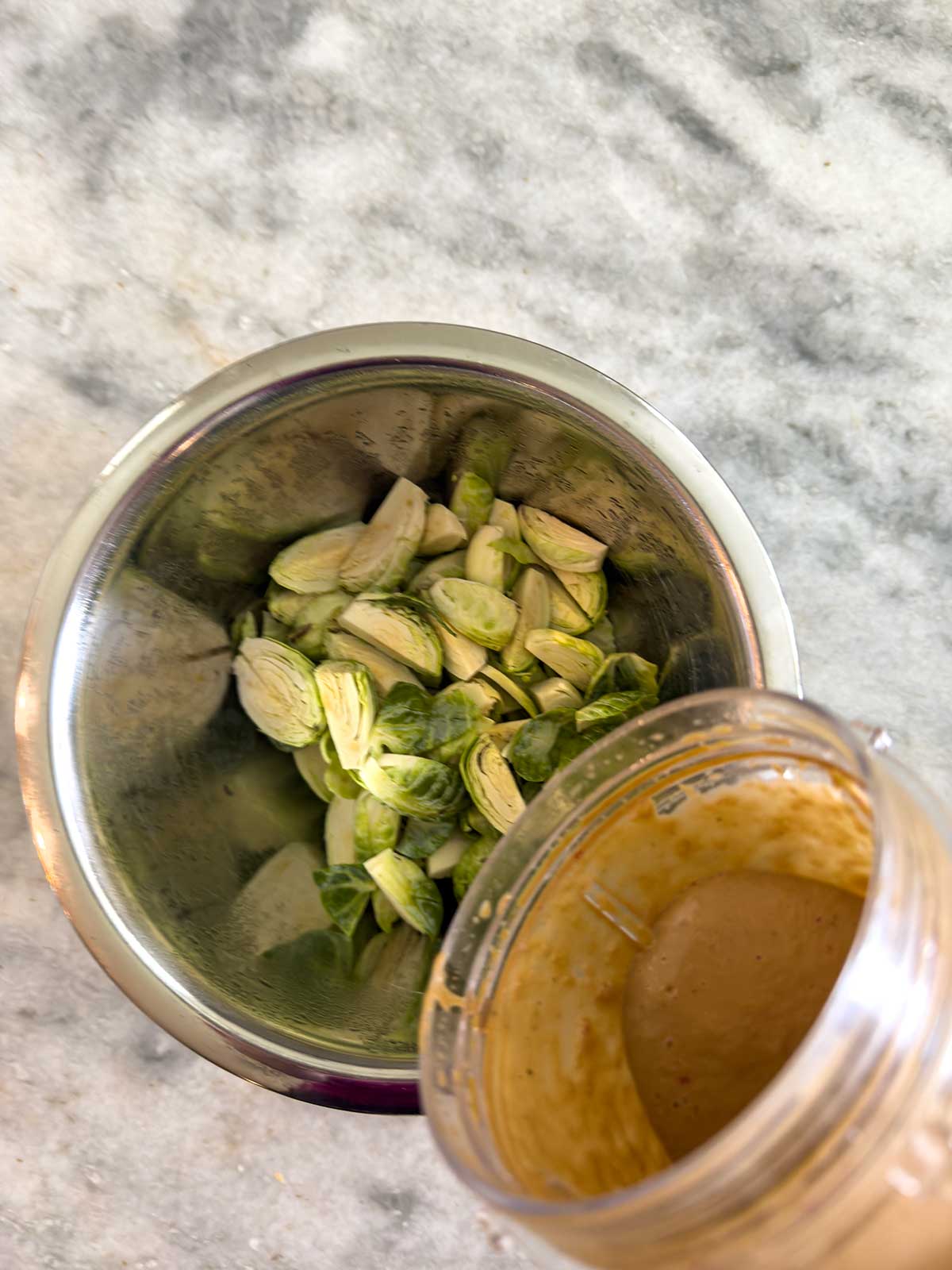 A metal bowl filled with chopped Brussels sprouts sits on a marble countertop next to a blender cup containing a creamy brown sauce.